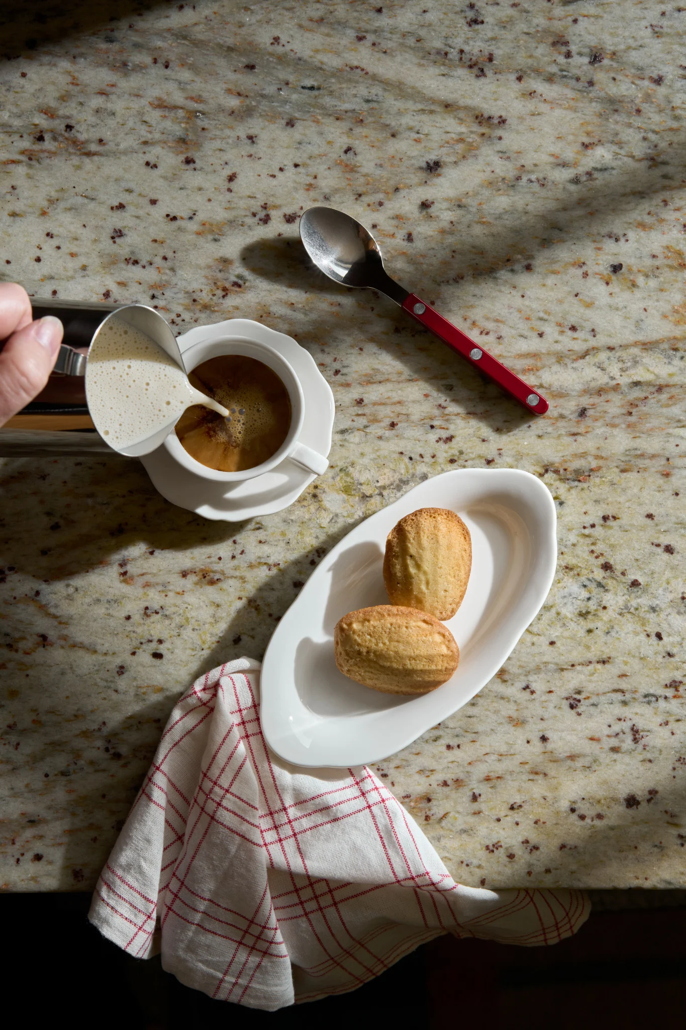Hånd heller skummet melk i kaffe. To madeleinekaker på en hvit tallerken og en skje med rødt håndtak hviler på en granittbenk.