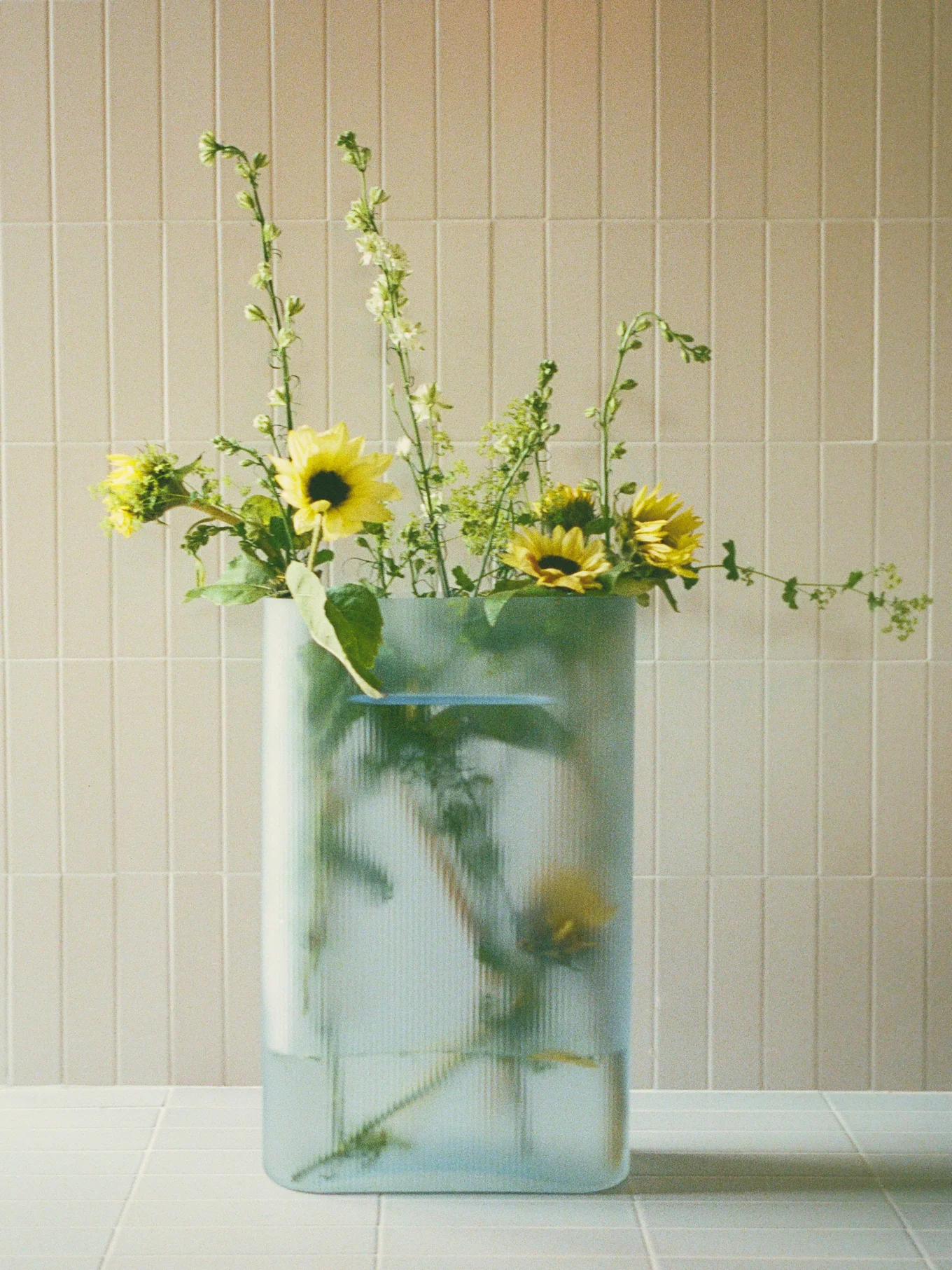Bukett med gule solsikker og grønne piggete blomster i en klar, riflet rektangulær vase på en flislagt overflate.