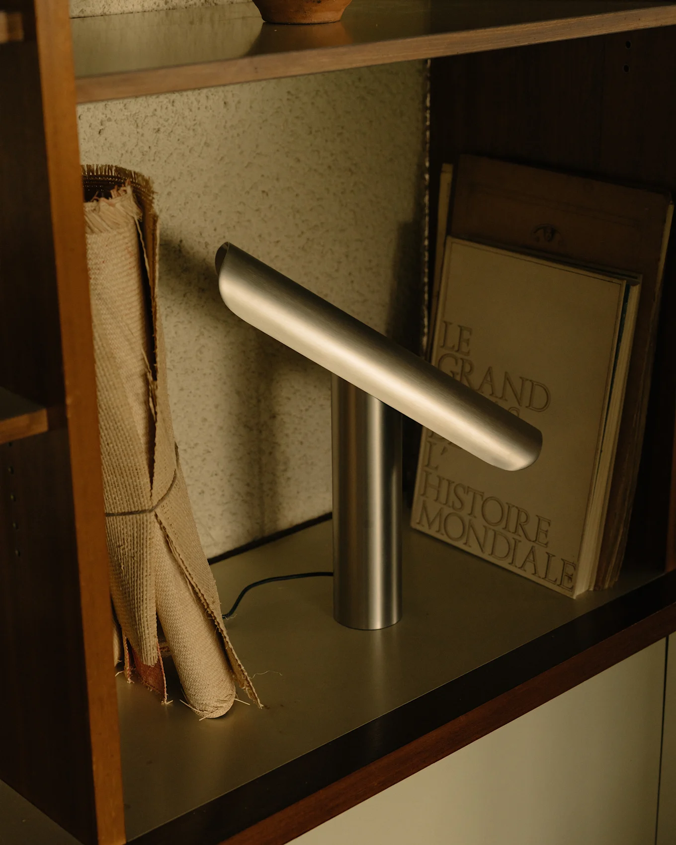 Moderne sølv skrivebordslampe med sylindrisk base og en vinklet horisontal lyslinje på en hylle.