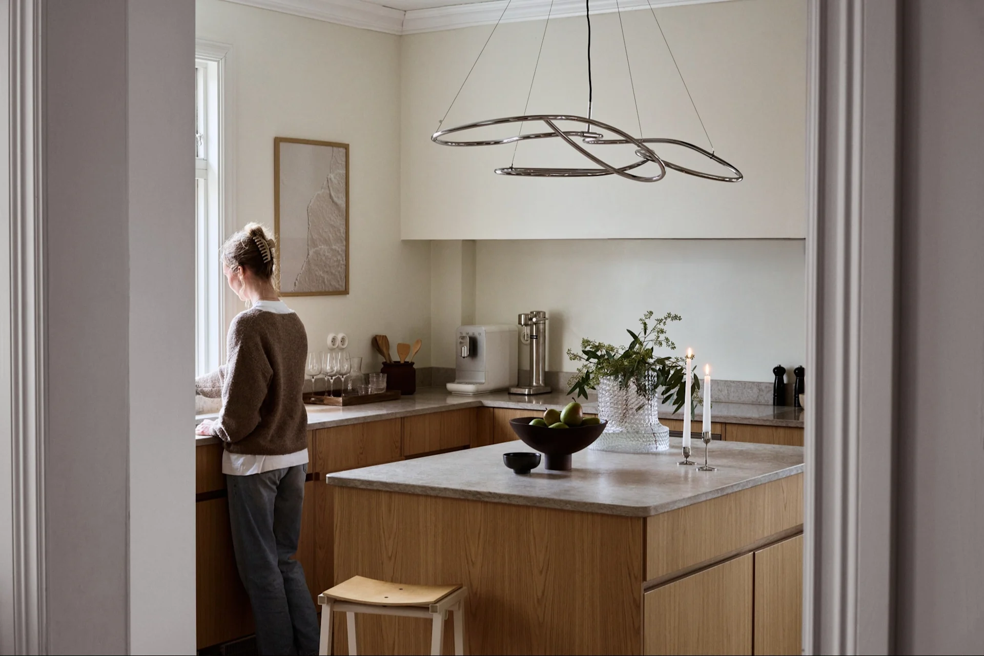 Kvinne ser ut av et vindu i et lyst, moderne kjøkken med lyse treskap, grå benkeplater og en skulpturell pendellampe.