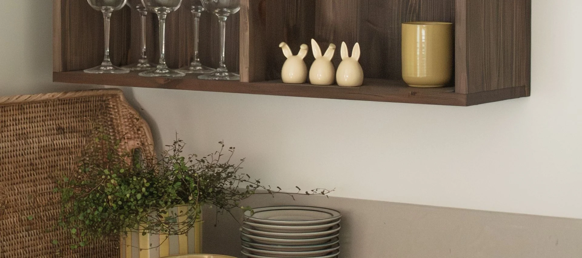 En trehylle med glass, tre kaninfigurer og et gult krus, med en flettet kurv og planter under.