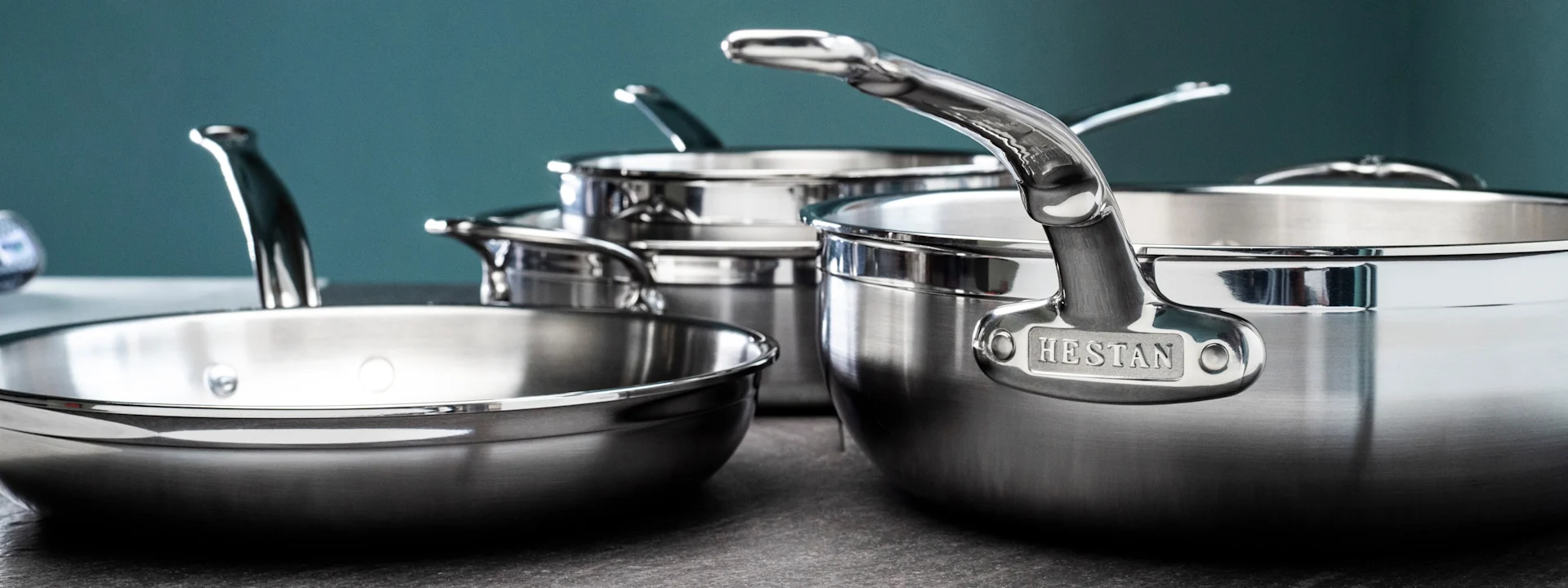 Skinnende gryter og panner i rustfritt stål, inkludert en stor Hestan-merket panne, på en mørk benkeplate.