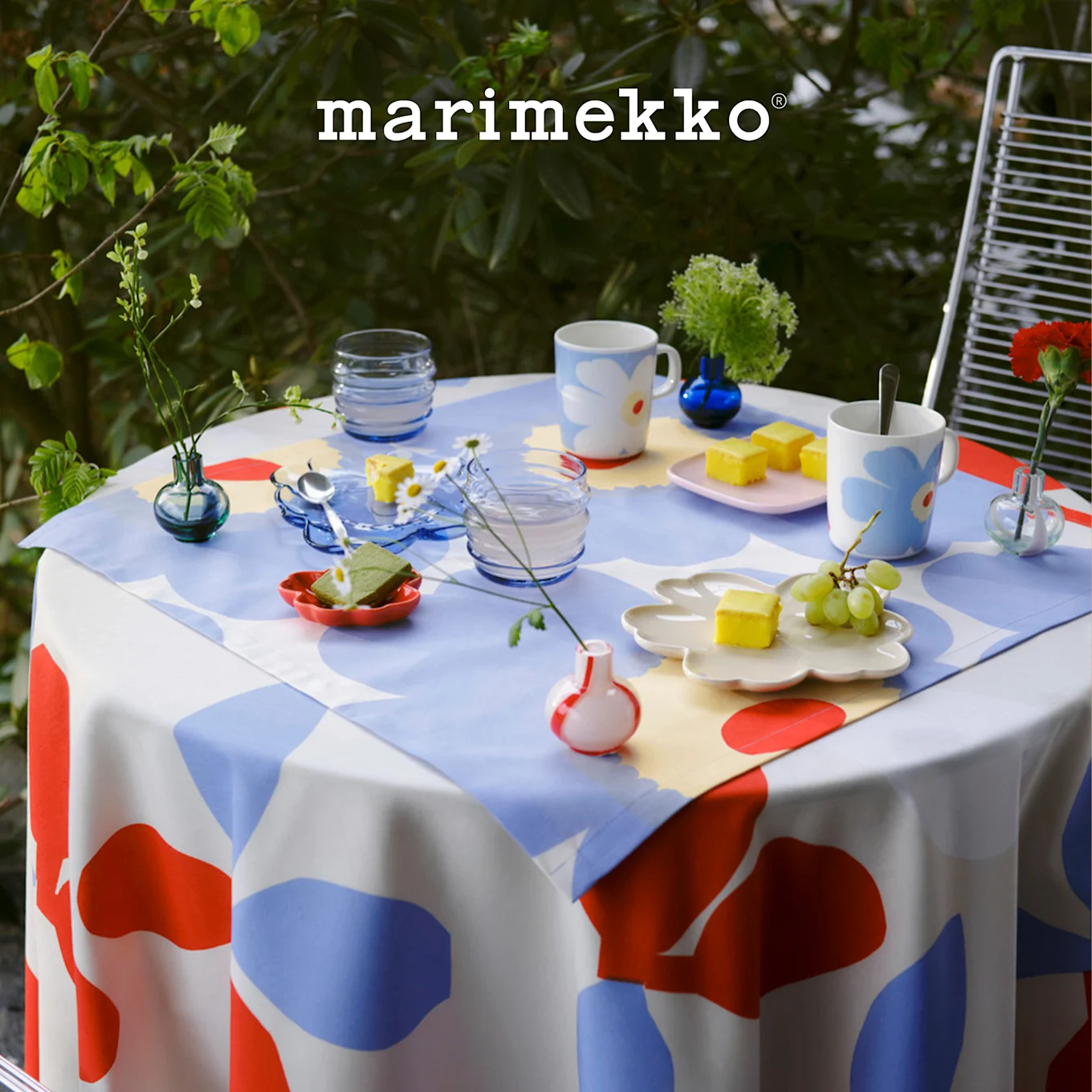 Utebord med Marimekko rød, blå, hvitt mønstret duk, dekket med blomstermugger, desserter og små planter. Marimekko logo.