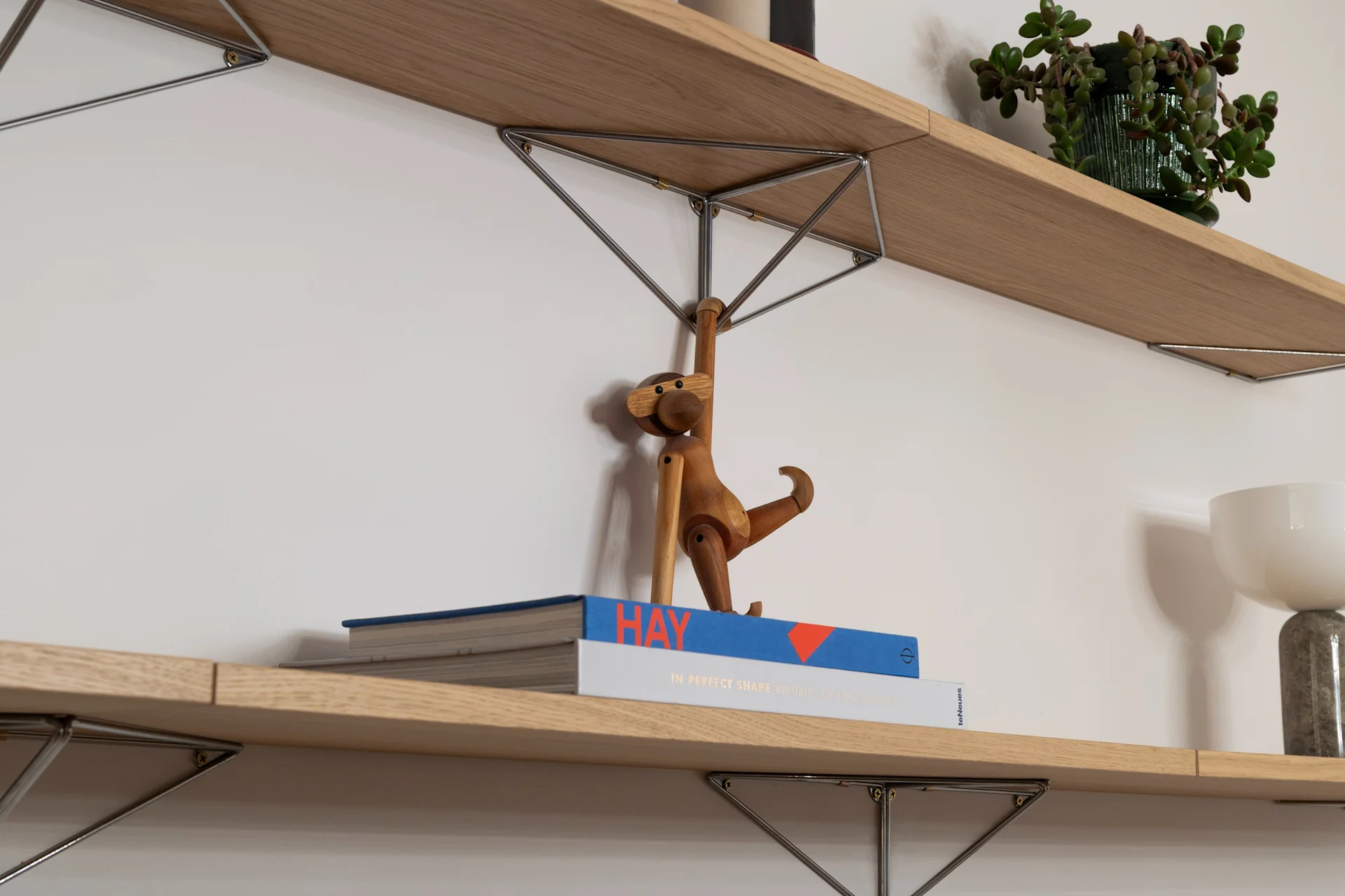 To trehyller på hvit vegg. Øverst: stripete lys, potteplante. Nederst: bøker, treape, hvit lampe.