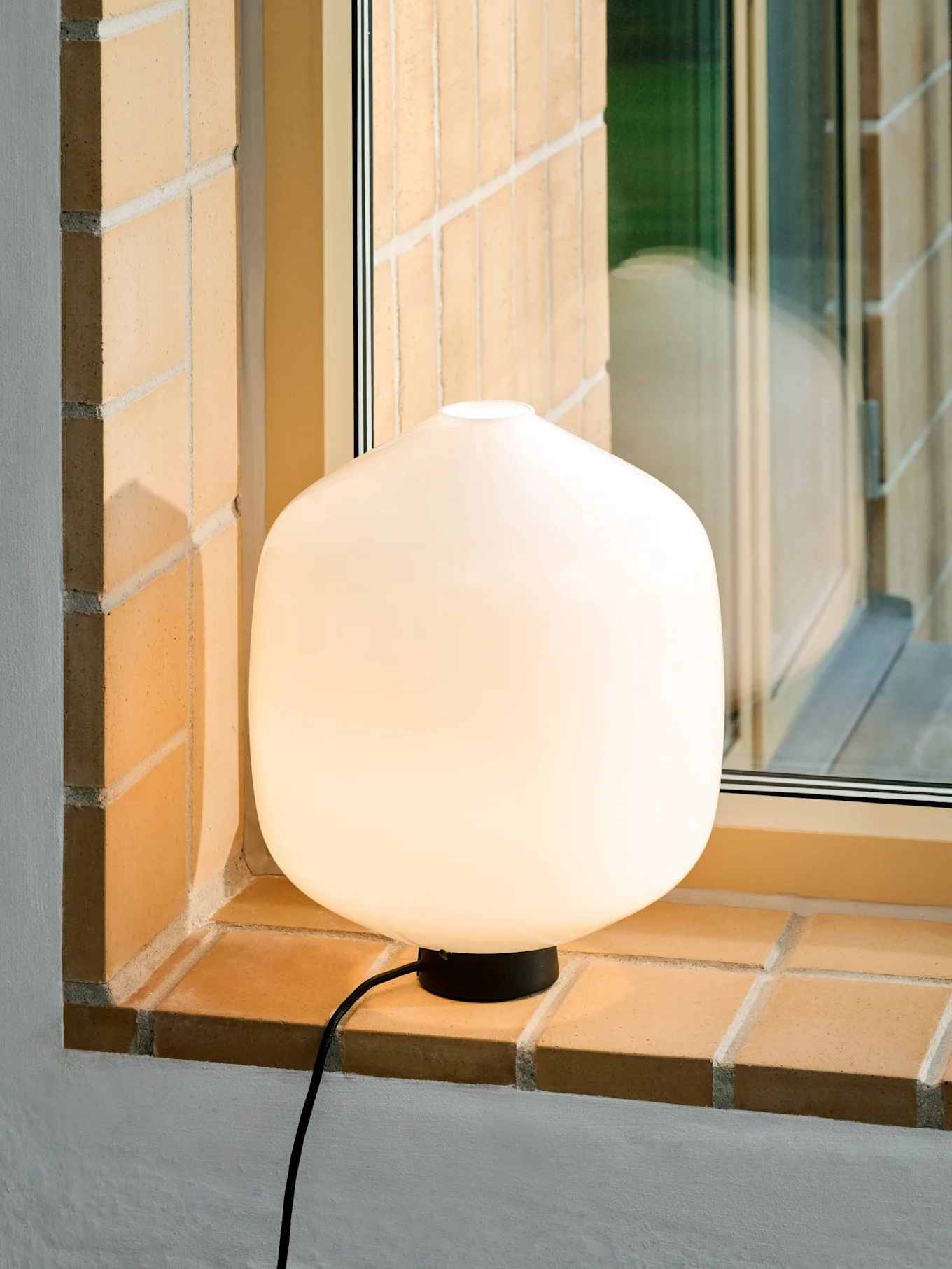 En glødende, avrundet hvit bordlampe med svart base og ledning på en mursteinsvinduskarm.