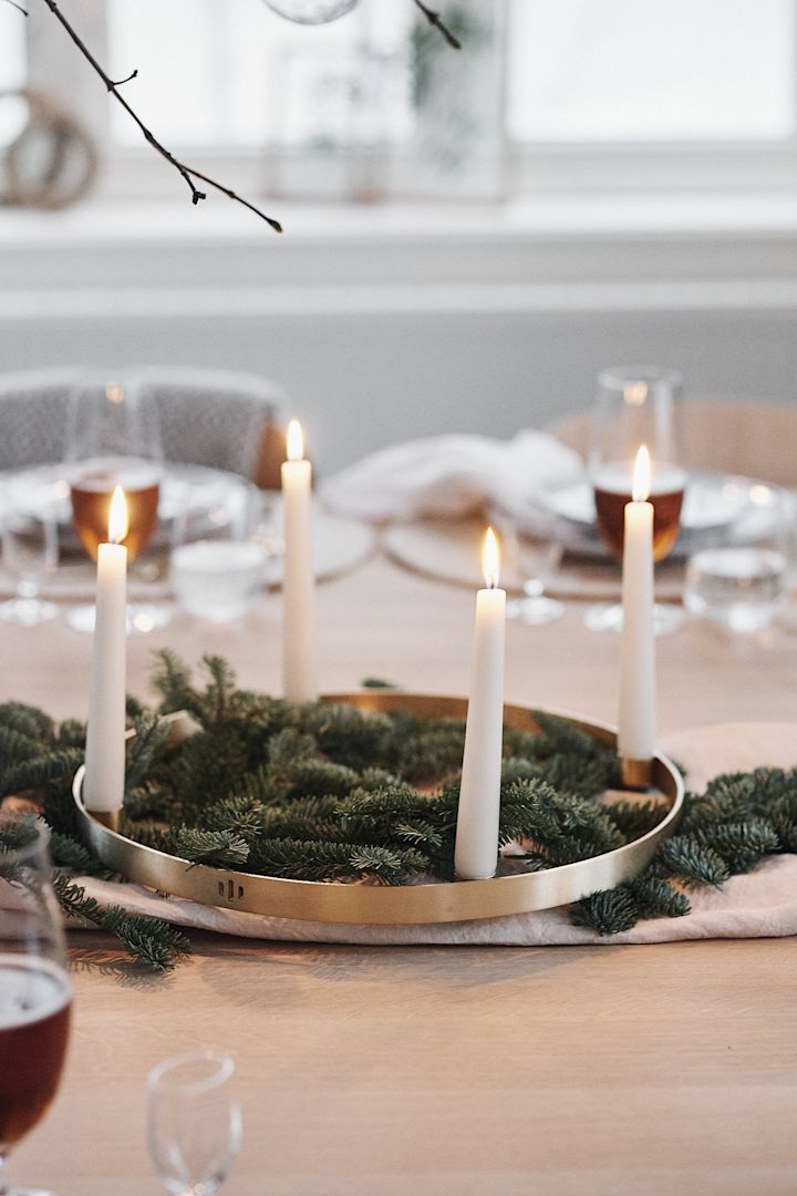 Granbar og en rund lysestake i messing med hvite lys står i midten av en hvit borddekking til jul og sprer stemning.