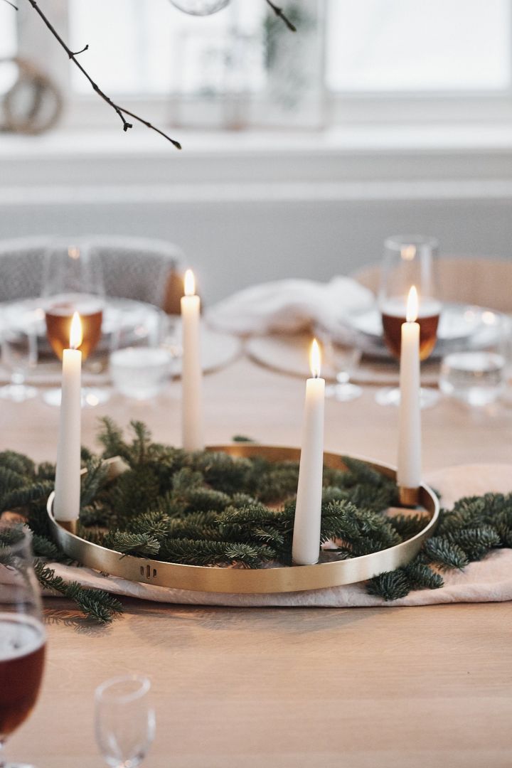 Granbar og en rund lysestake i messing med hvite lys står i midten av en hvit borddekking til jul og sprer stemning.
