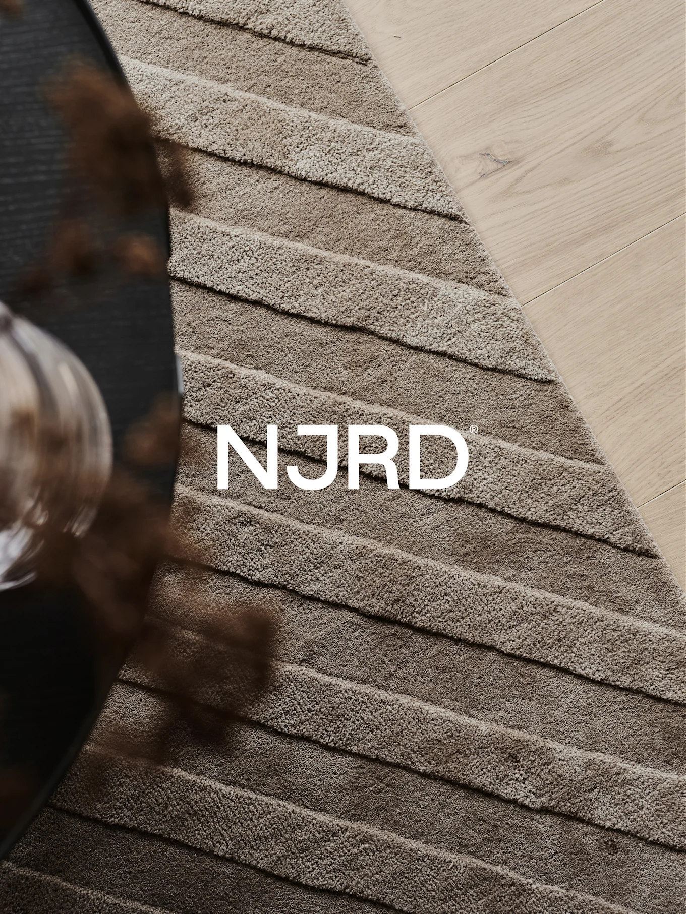 Nærbilde av et lysebrunt rya-teppe med horisontale riller, delvis på et lyst tregulv, med den hvite 'NJRD'-logoen.