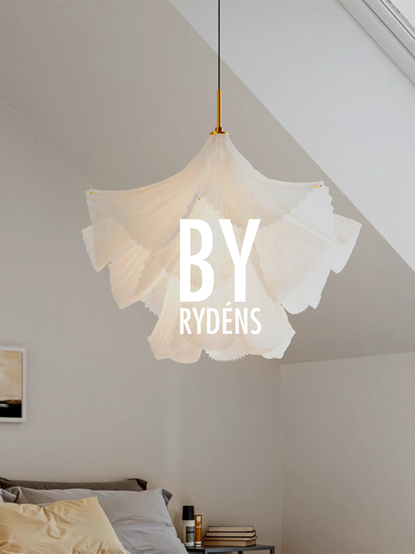 En hvit, plissert skulpturell pendellampe henger over en seng med puter og et nattbord. 'BY RYDÉNS' er lagt over lampen.