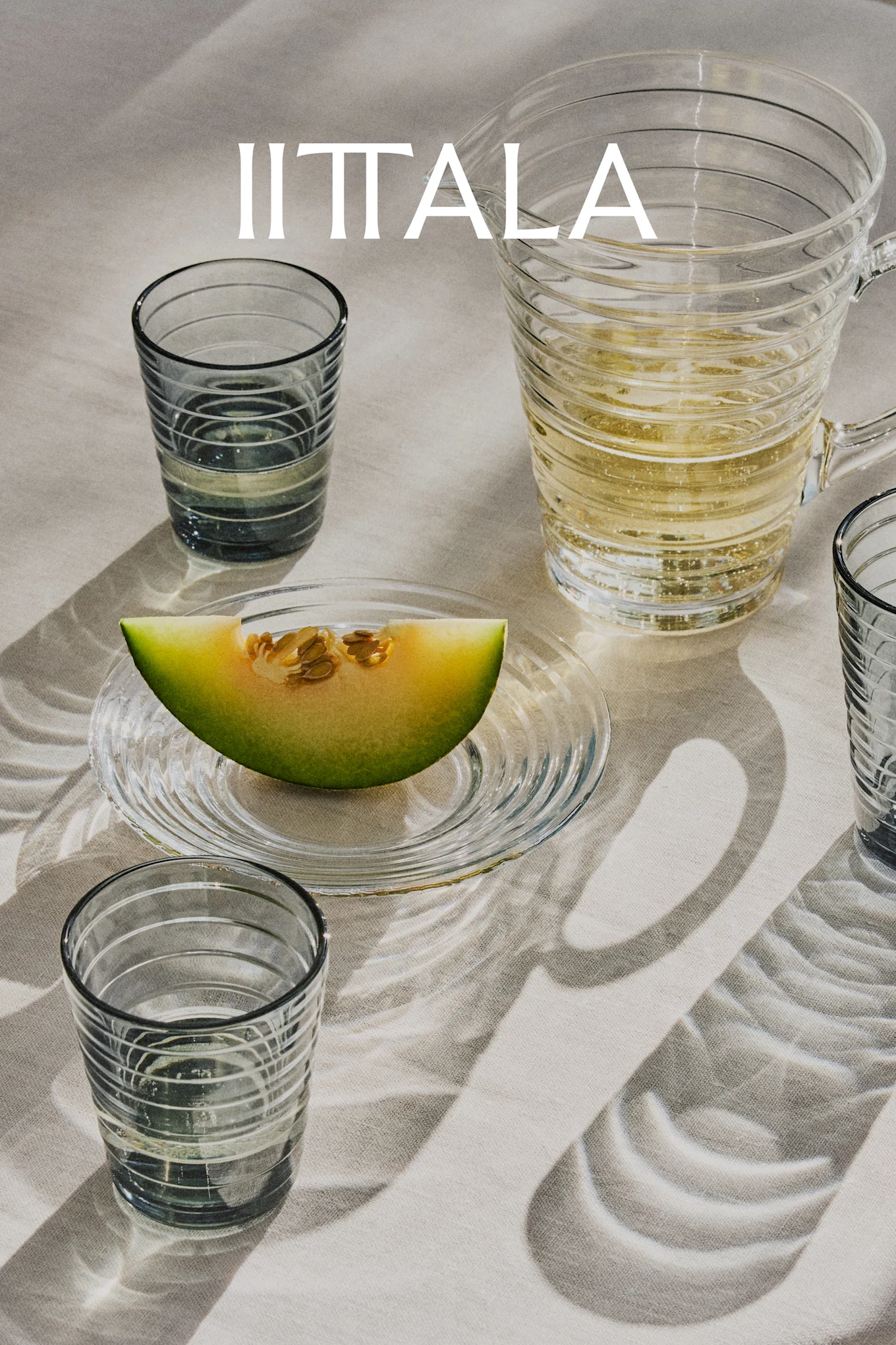 IITTALA-logo over glass, en mugge med musserende drikke og en melonskive på tallerken, alt på lyst stoff.