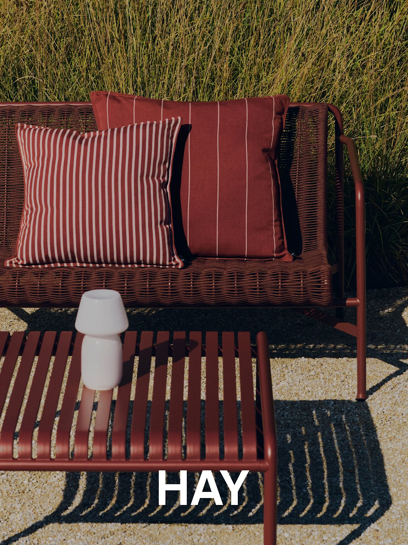 Utendørsscene med en vevd rød sofa, stripete puter, et lamellert salongbord med en hvit lampe og høyt gress i bakgrunnen. HAY-logo.