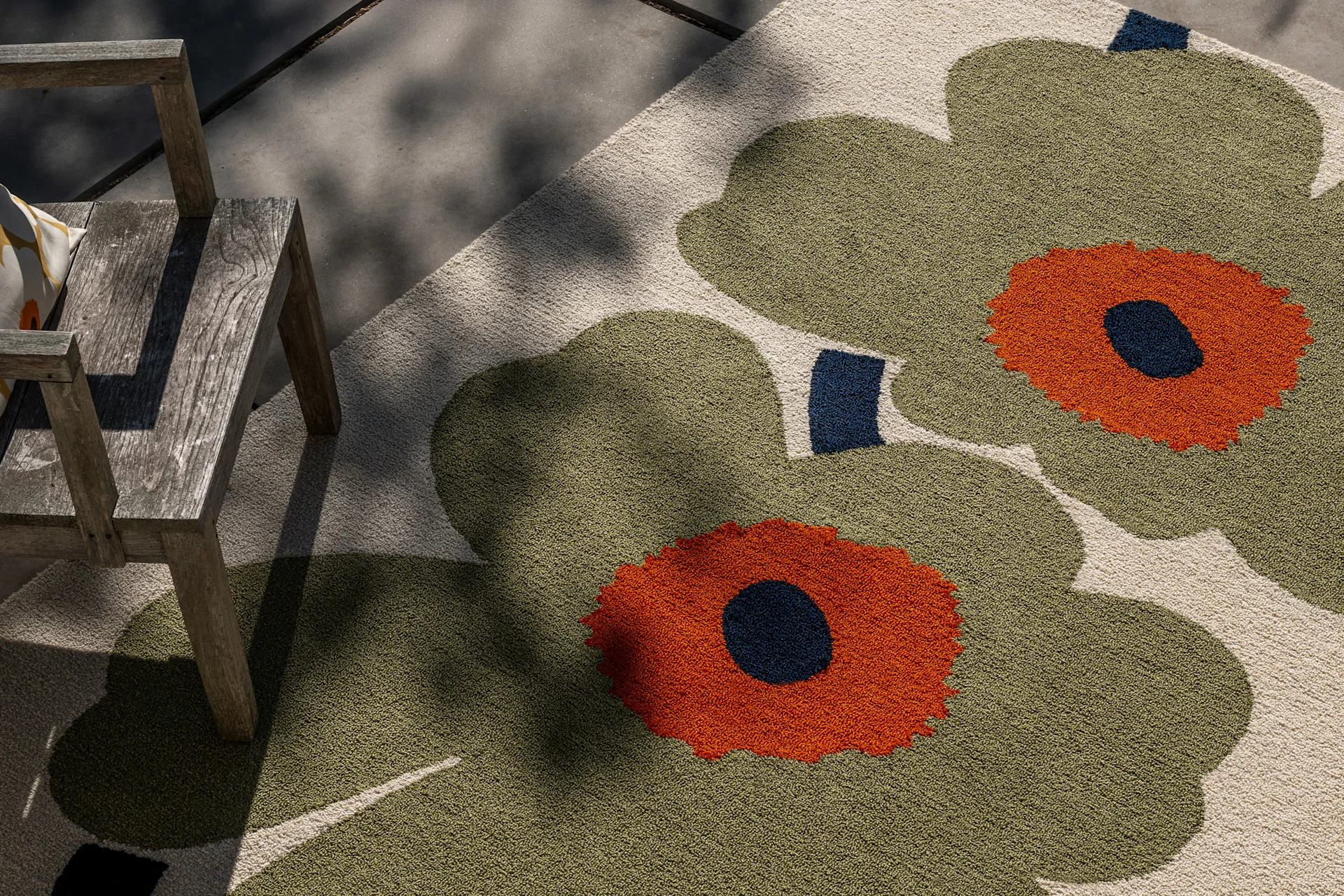 Utendørs teppe med stort oliven grønt, oransje og mørkeblått blomstermønster, ved siden av en rustikk trestol i flekkete sollys.
