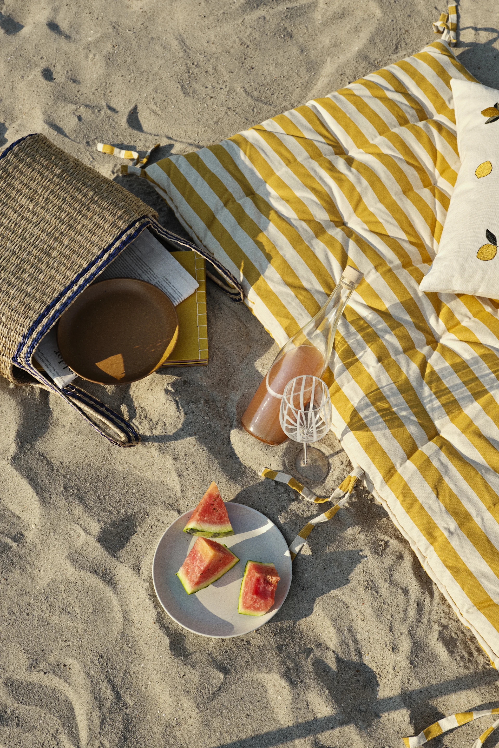 Strandpiknik med gulstripet matte, stråveske, vannmelon, drikkeflaske, mønstret glass og sitronpute.