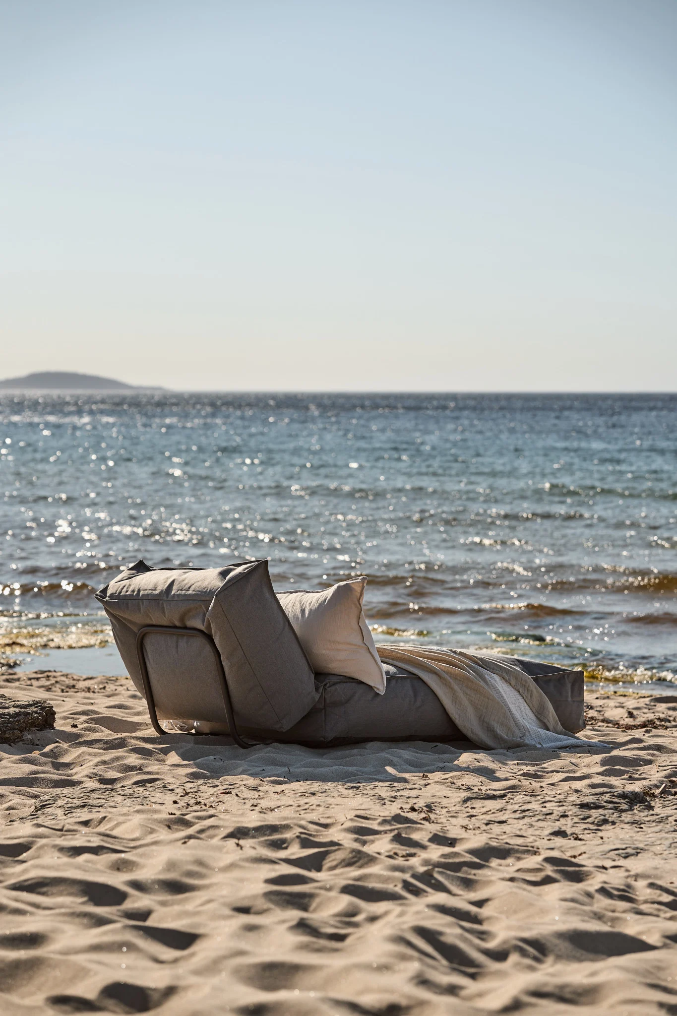 Grå solseng med hvit pute og lyst pledd på en sandstrand ved sjøen.