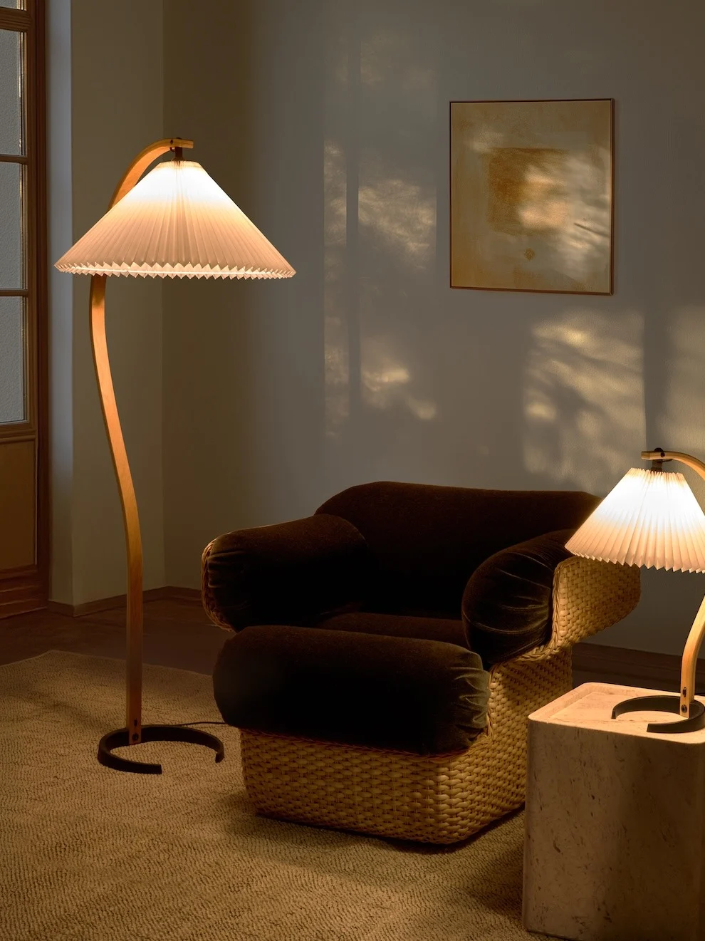 Koselig rom med en buet gulvlampe, mørk lenestol med flettet base og en mindre bordlampe. Varmt lys skaper skygger.