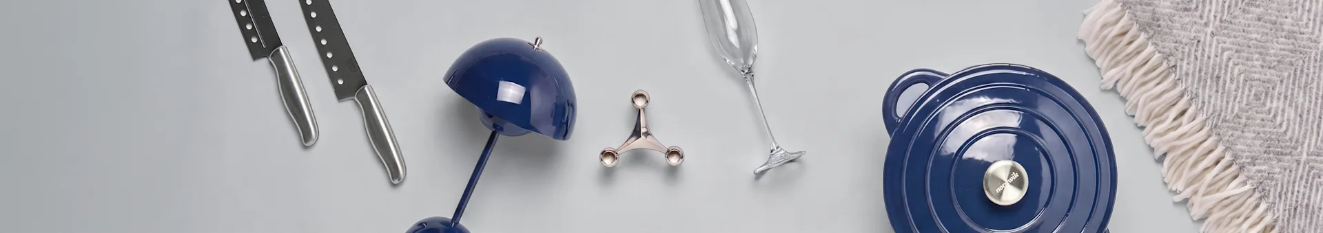 To kniver, en blå lampe, en sølvholder, et champagneglass, en blå gryte og et grått mønstret teppe på en lys overflate.
