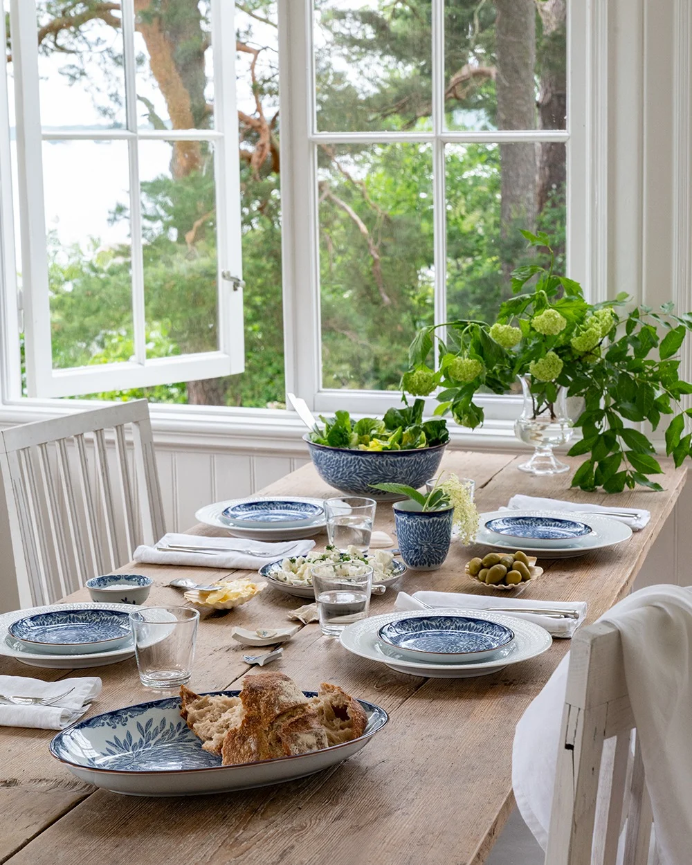 Et rustikt spisebord i tre dekket med blå og hvite servise, salat, brød og oliven, ved et åpent vindu med utsikt over grønne trær og vann.