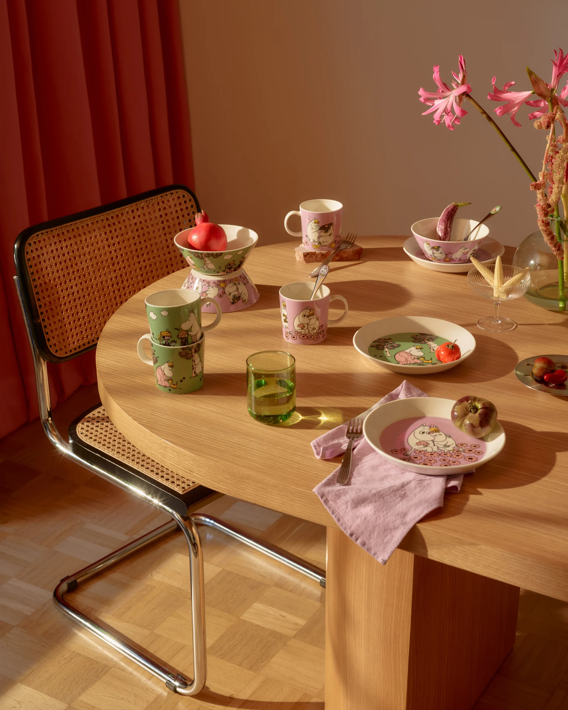 Spisebord med rosa og grønt Mummi-servise, friske frukter, et grønt glass og rosa blomster.