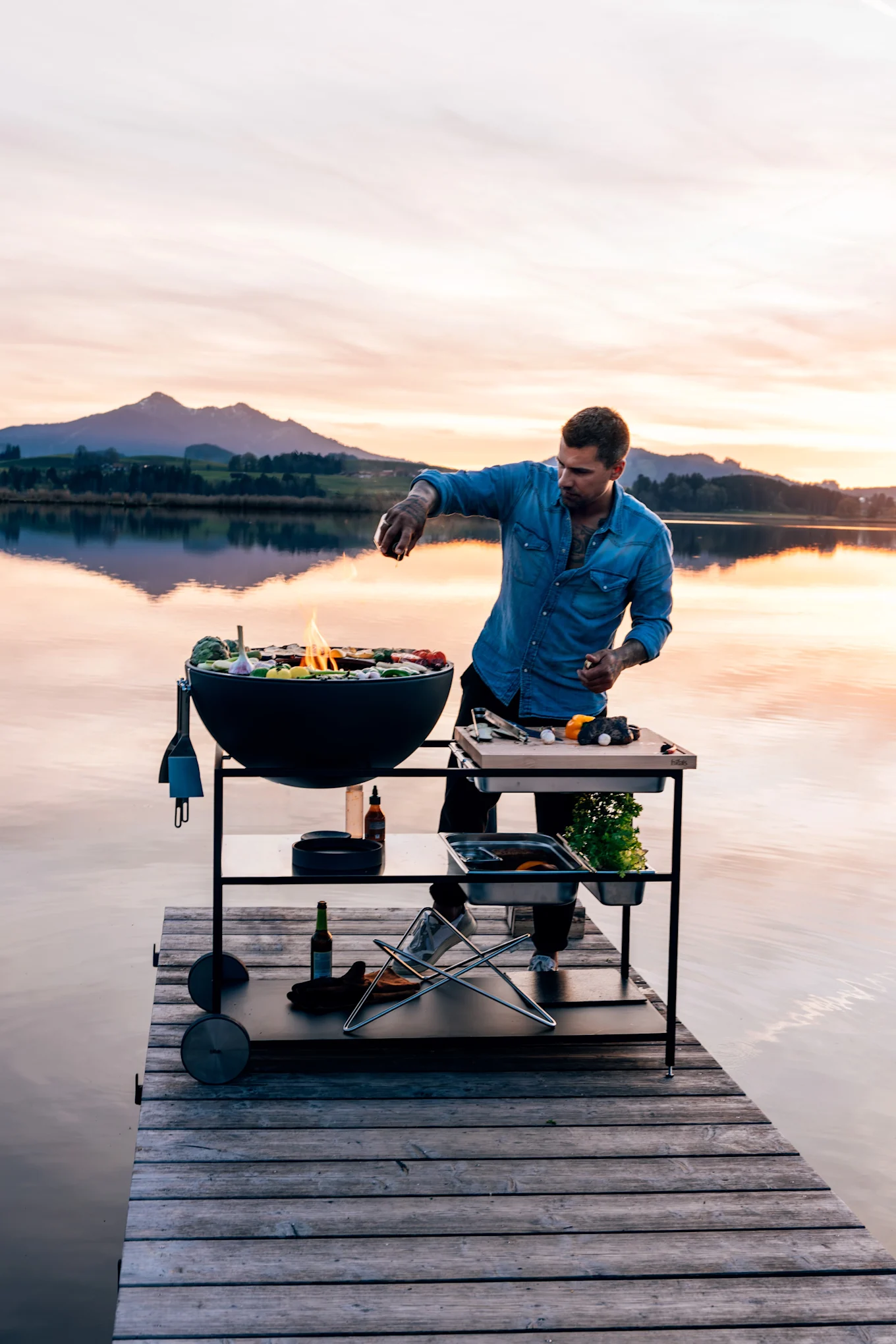 Mann i dongeriskjorte griller mat på en rund svart grill og forberedelsesbord på en trebrygge ved solnedgang.