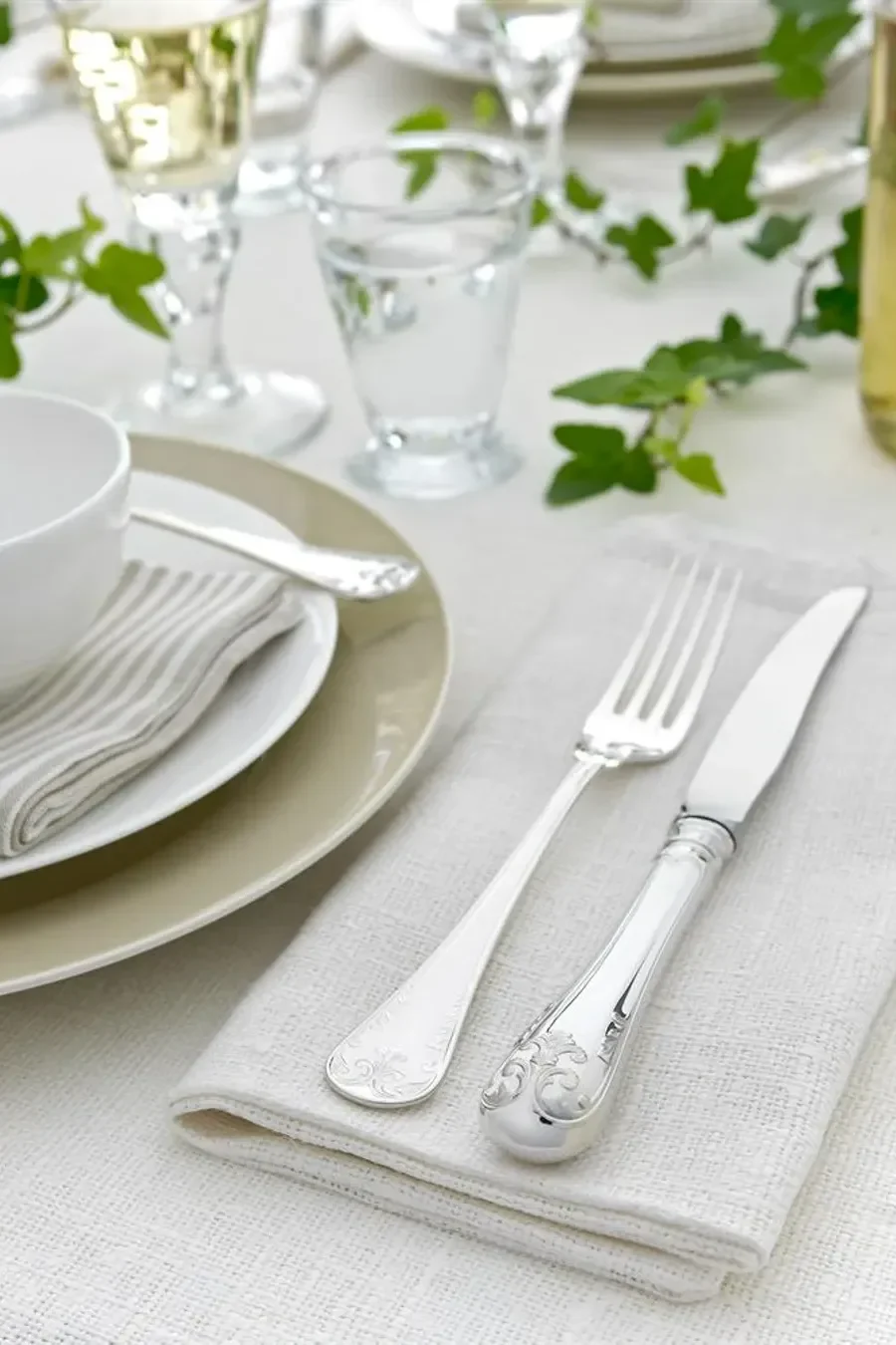 Elegant borddekking med utsmykket sølvbestikk, stablede tallerkener, glass og grønn eføy på hvit duk.