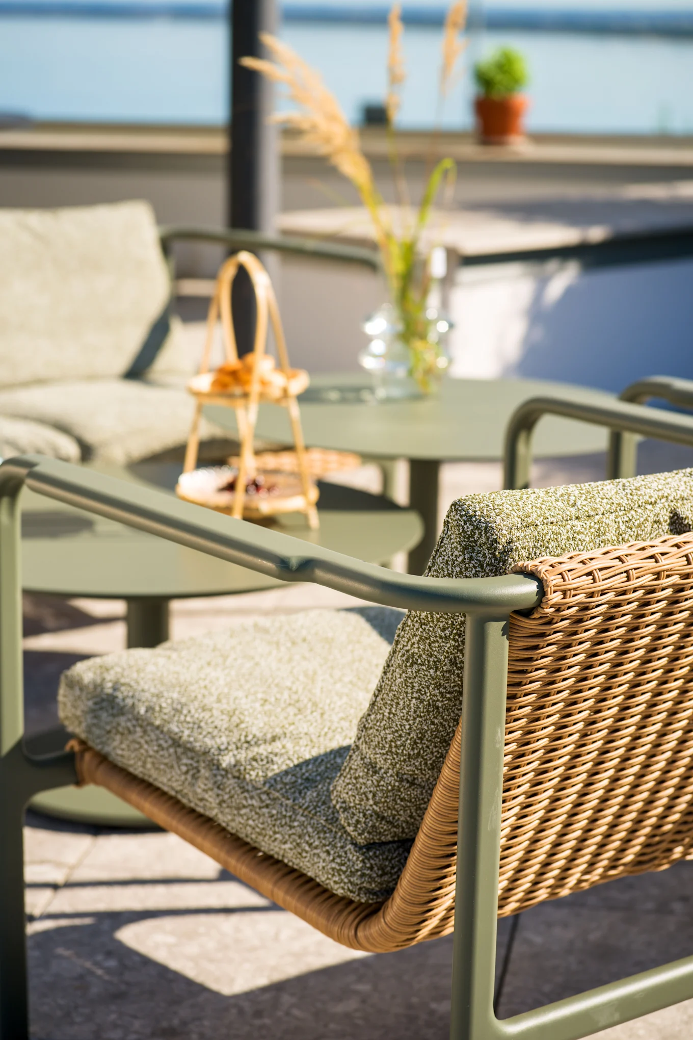 Utendørs terrassestol med olivengrønn ramme, flettet rygg i rotting og grønne puter. Matchende bord og stol i uskarp bakgrunn nær vann.