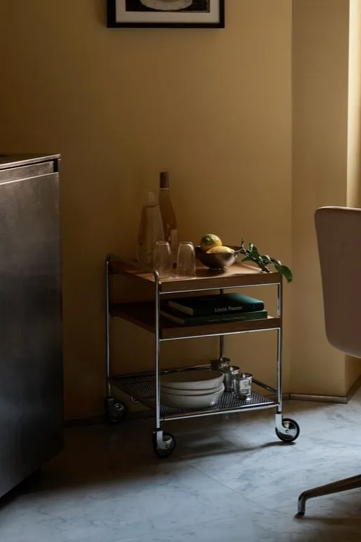 En treroms tralle med metallramme og trehyller, som inneholder flasker, glass, sitroner, bøker og tallerkener.