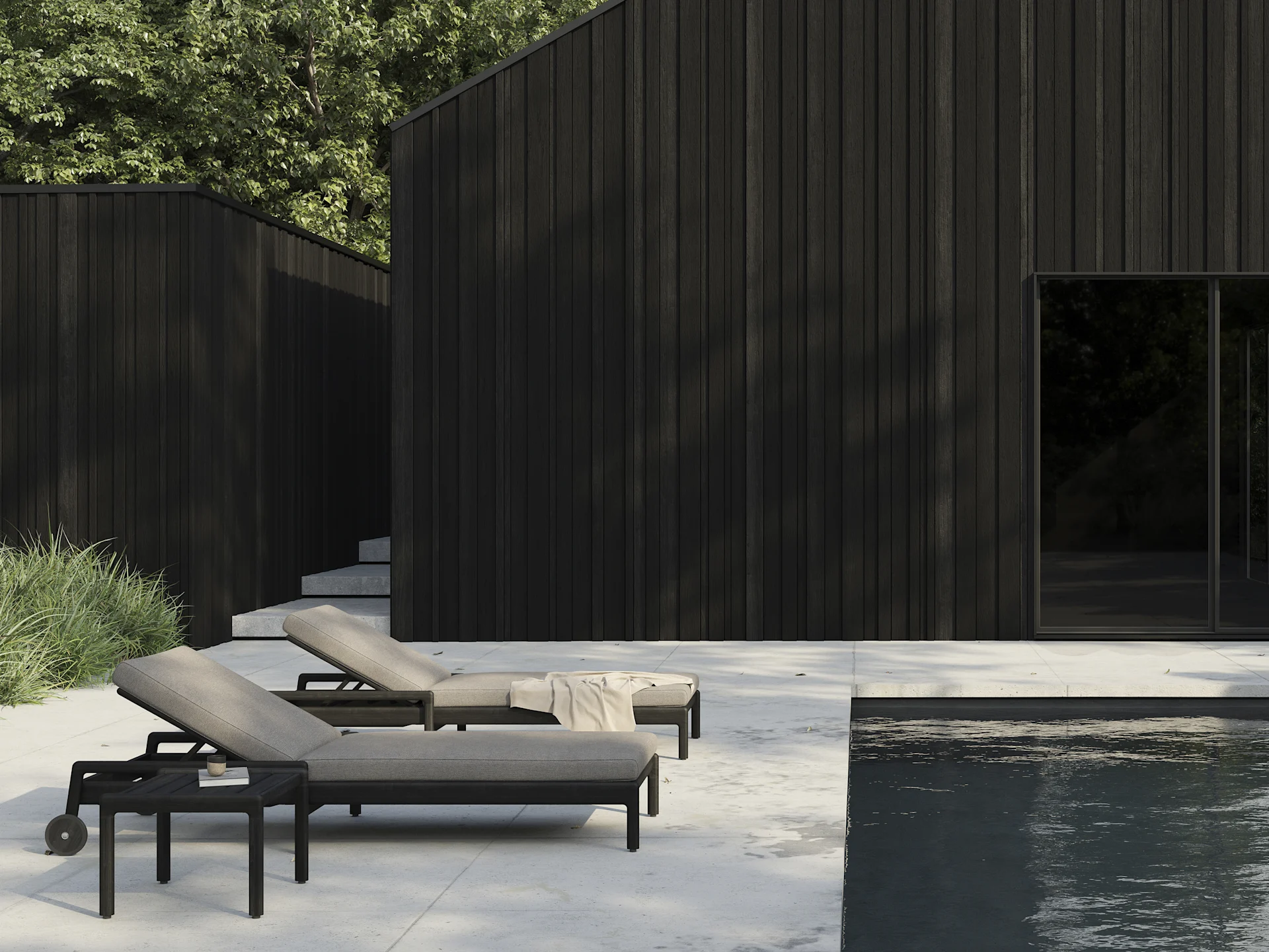 To mørke solsenger med lyse puter på en uteplass ved et svømmebasseng, med moderne mørke trebygninger i bakgrunnen.