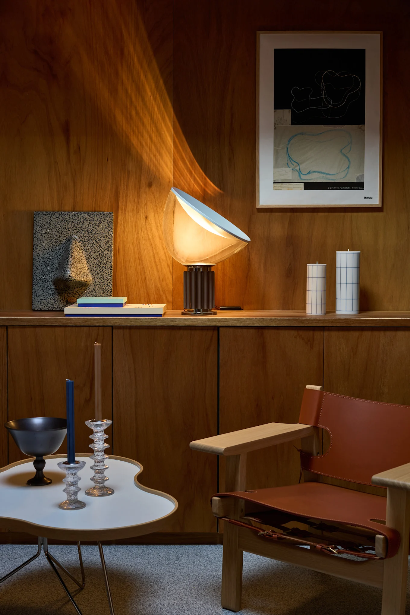 Et oppholdsrom med trevegger, en glødende bordlampe, abstrakt veggkunst, dekorative lys, et moderne sidebord og en skinnlenestol.