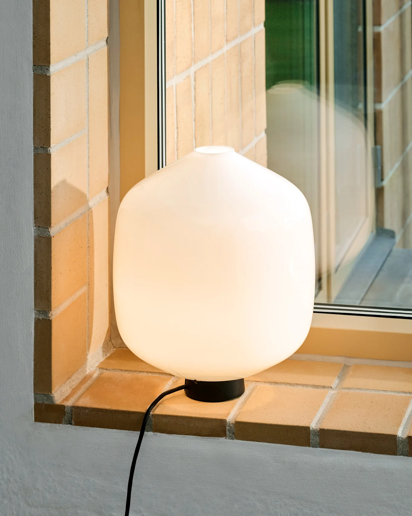 En glødende hvit, avrundet bordlampe med svart base og ledning står på en lys mursteinsvinduskarm.