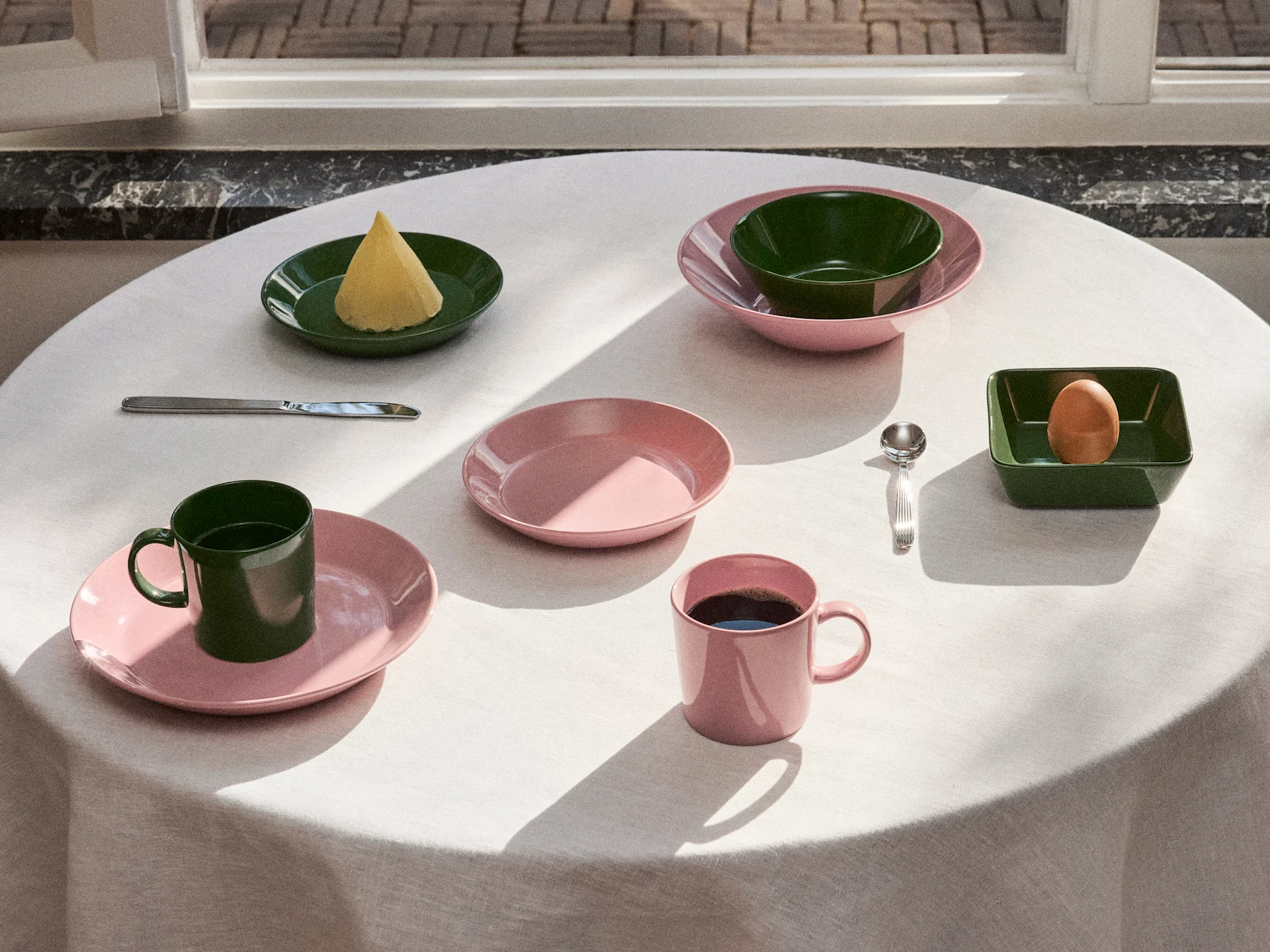 Bord dekket med rosa og mørkegrønt servise, tallerkener, skåler og krus, med smør, et egg og kaffe i naturlig lys.