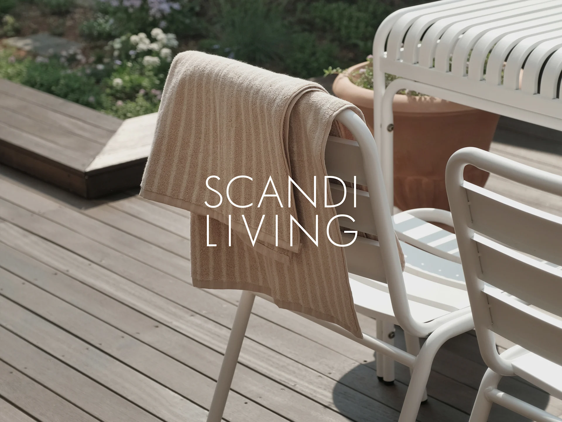 Et beige stripet håndkle henger på en hvit utendørs stol på en treterrasse med planter. Tekst: SCANDI LIVING.