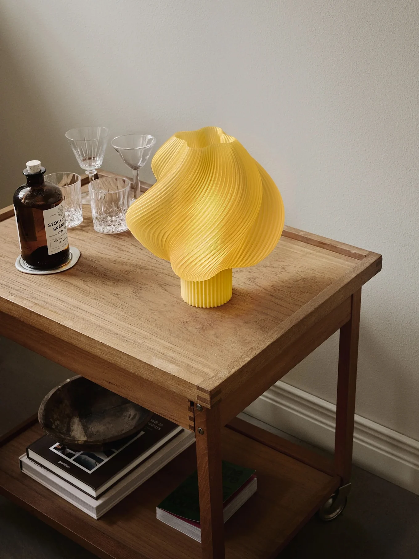 Gul plissert lampe på en rullende trekasse med en ginflaske, glass og bøker på nederste hylle.
