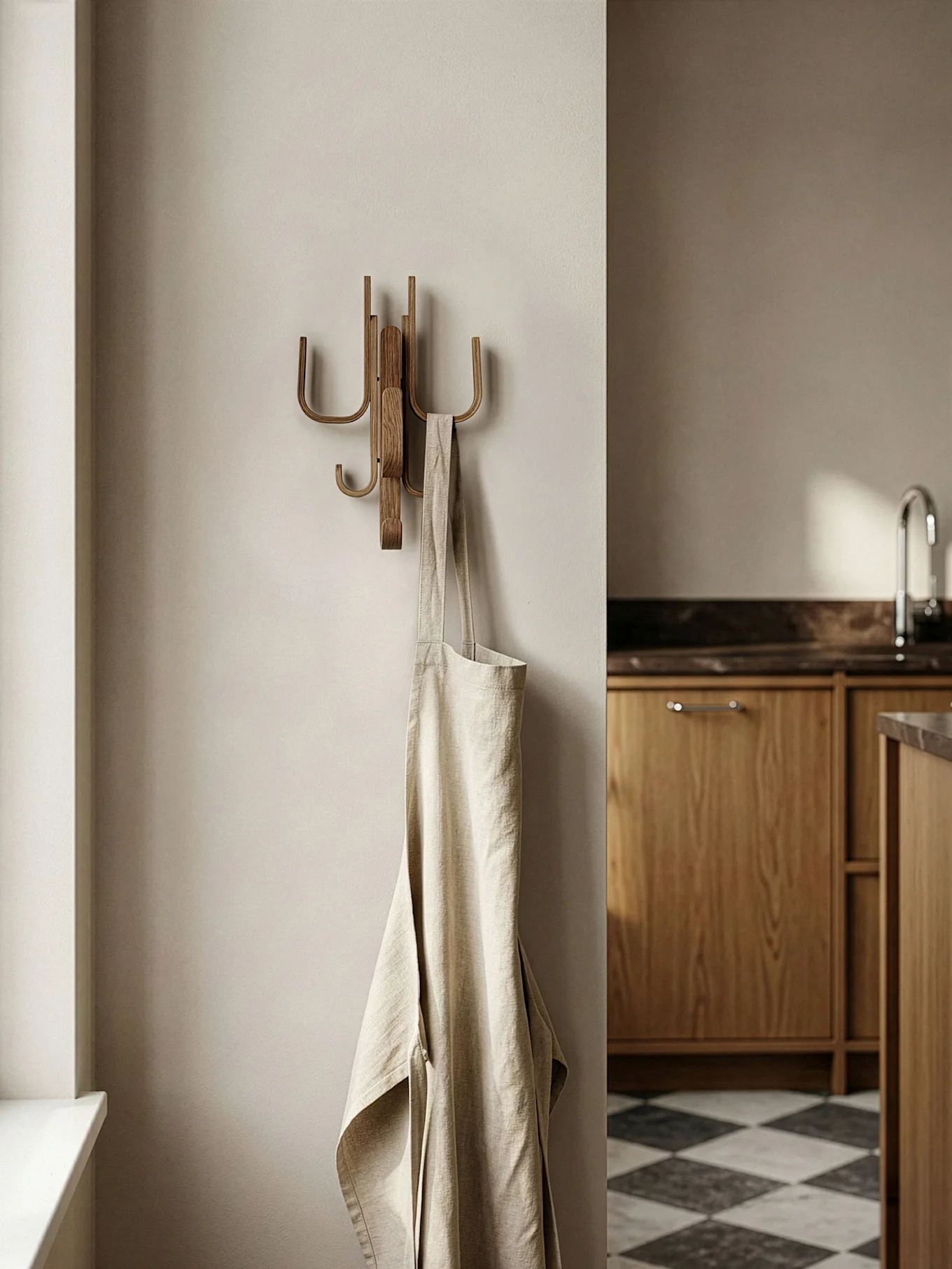 Et lyst beige linforkle henger på en veggkrok i tre på et moderne kjøkken med treskap og rutete gulv.