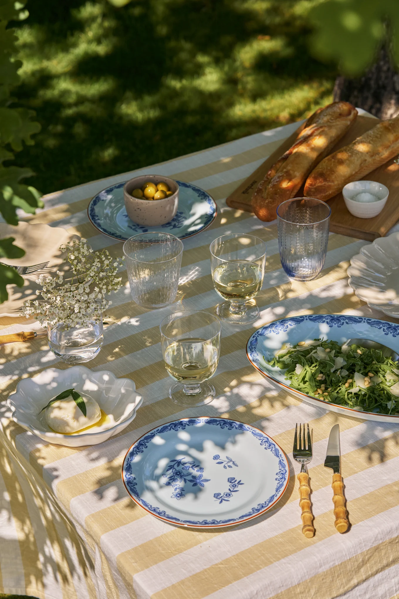 Utendørs spisebord dekket med hvit- og gulstripet duk, blå blomsterplater, vin, salat, mozzarella, oliven og baguetter.