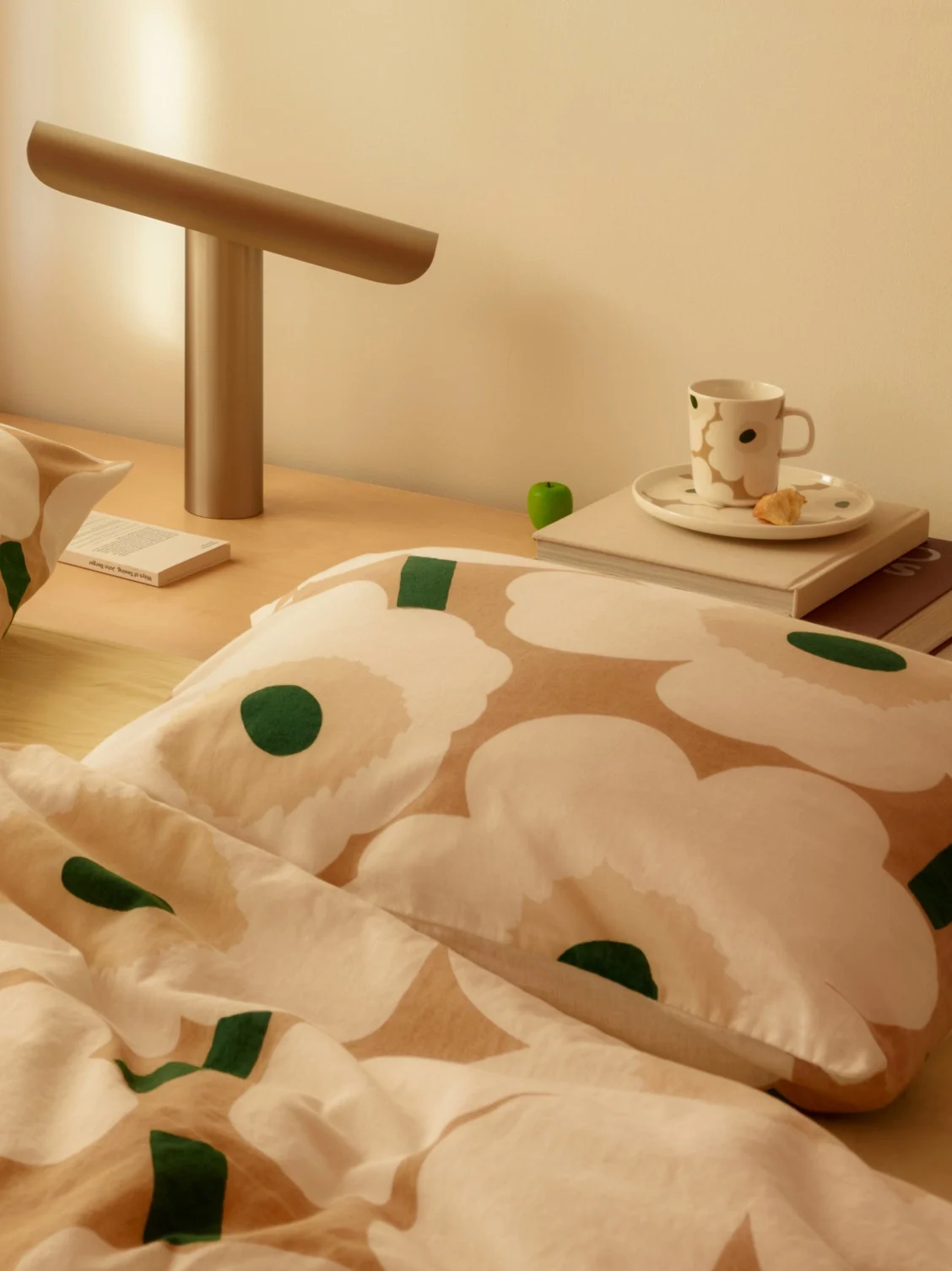 Sengekantsmotiv med hvit og beige blomstret sengetøy, en moderne lampe, en kopp og en bok på et nattbord i tre.