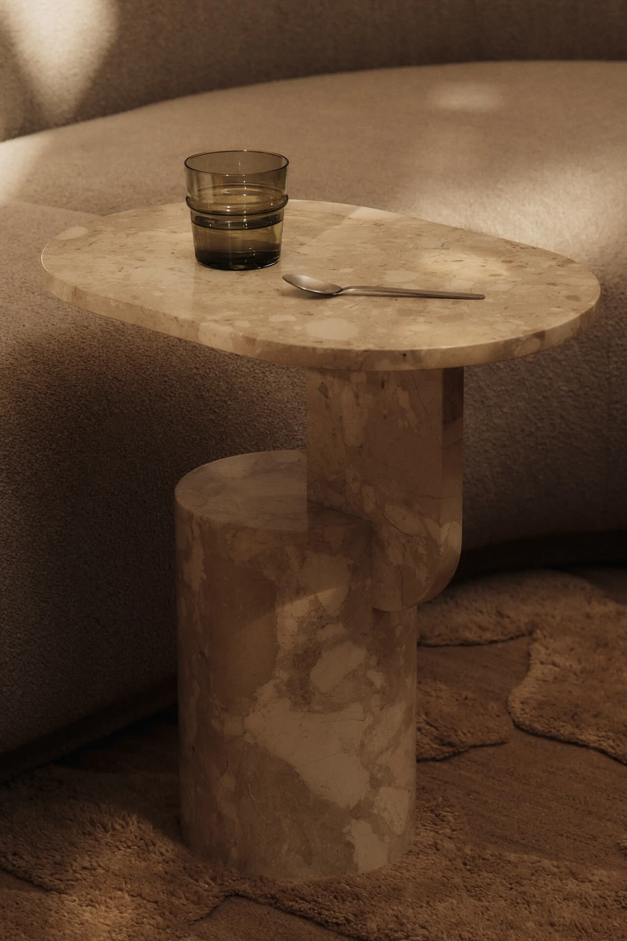 Beige marmor sidebord med et mørkegrønt glass vann og en sølvskje, ved siden av en teksturert sofa og et teppe.