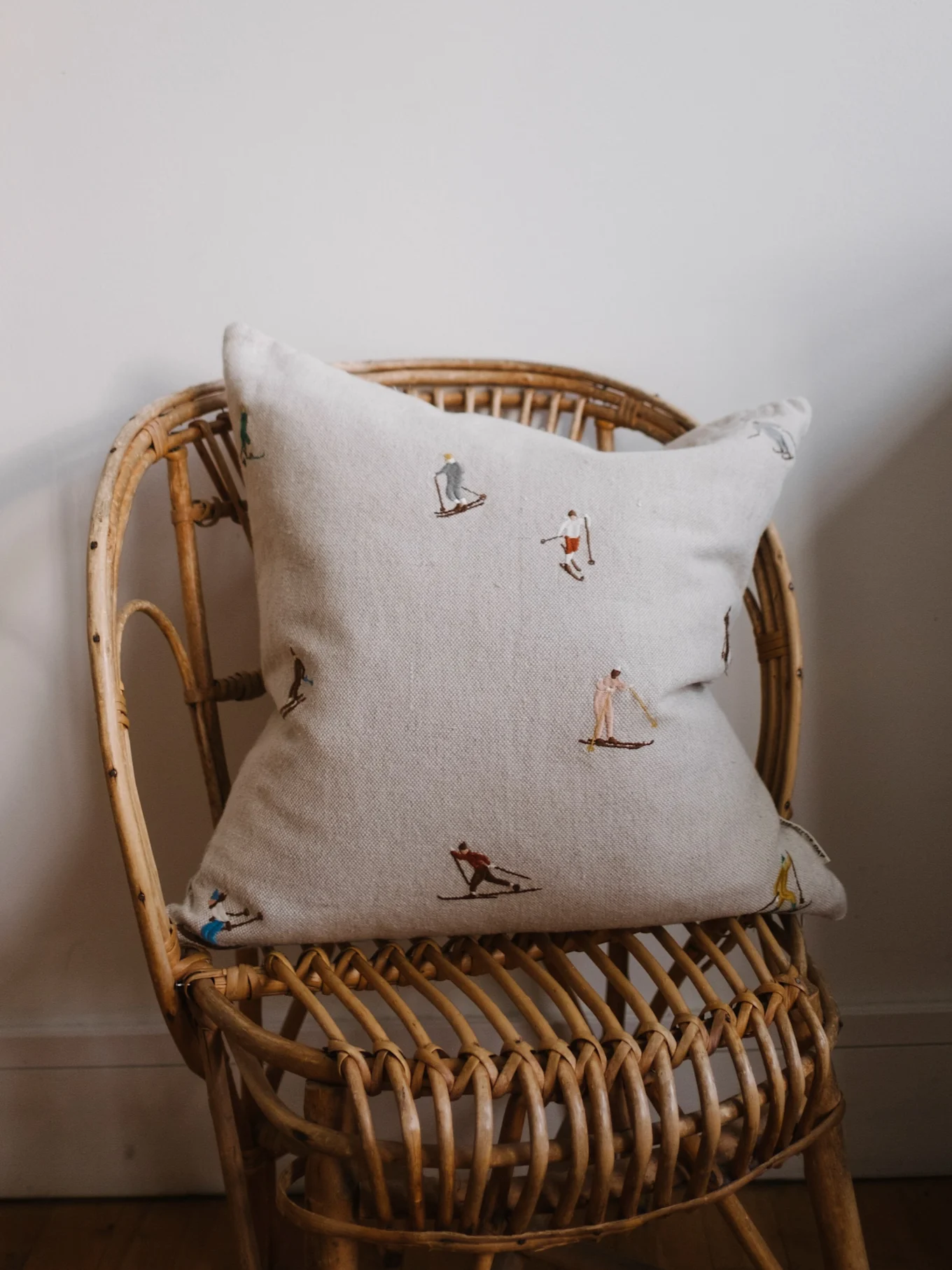 Lys beige pute med broderte skifigurer på en rottingstol mot en off-white vegg.
