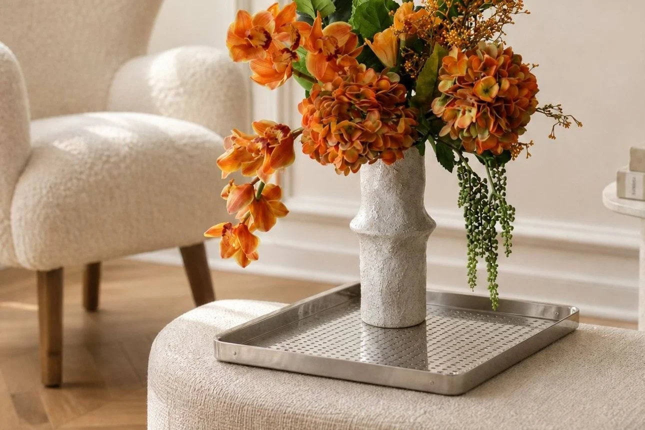 Levende oransje blomster i en teksturert hvit vase på et sølvbrett, plassert på en lys ottoman i en moderne stue.