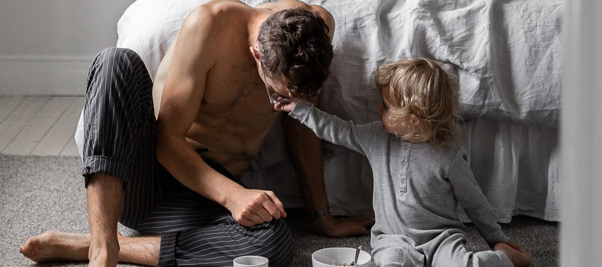 Far og barn sitter på gulvet i soverommet, barnet leker med fars briller, frokostvarer i nærheten.