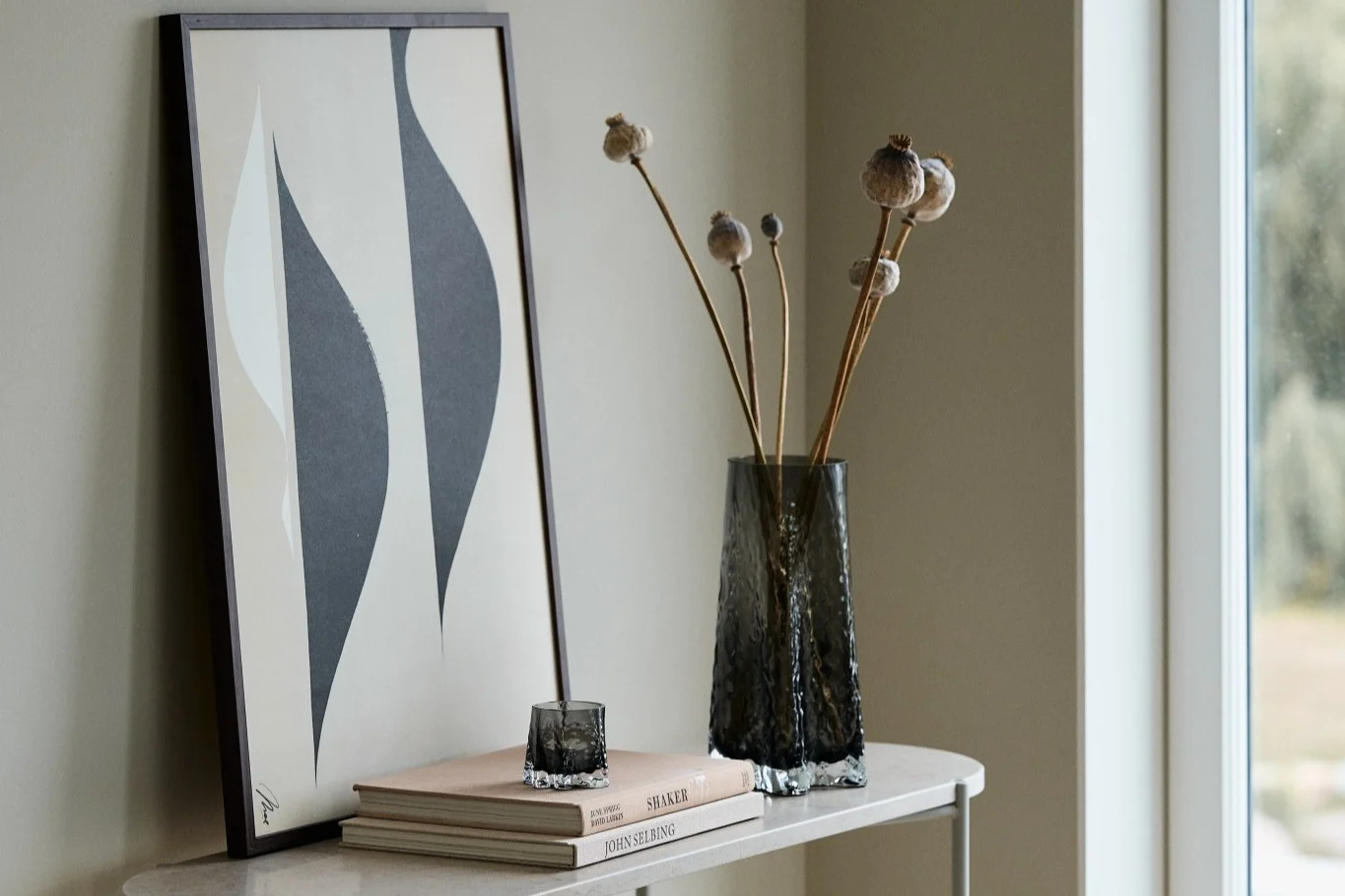 Her ser du Gry-vasen 30cm og Gry-telysholder begge i fargen Smoke fra Cooee Design stående på et sidebord foran et trykk.