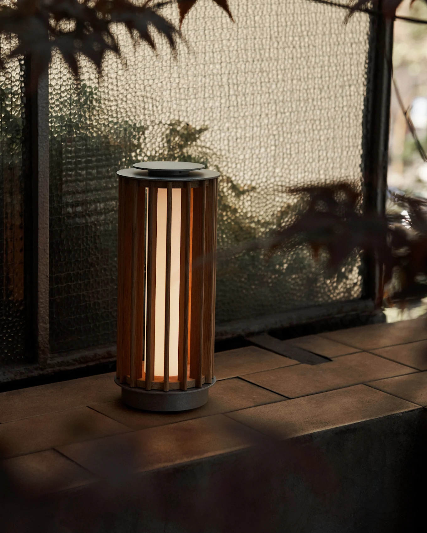En sylindrisk bærbar lampe med lamellert trekropp og varm glød, på en flislagt overflate ved et teksturert glassvindu.