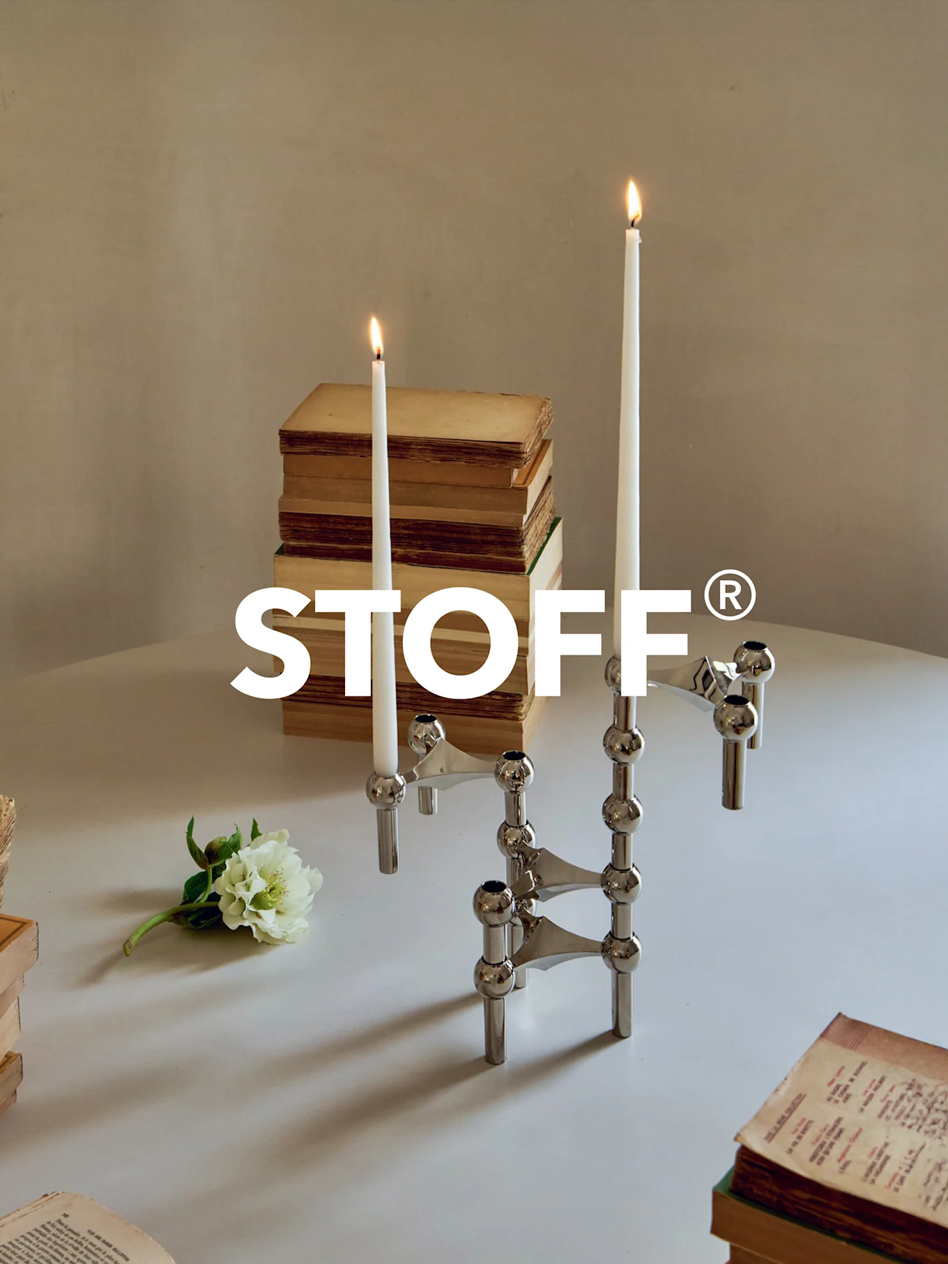 Polert krom modulær lysestake med to tente lys, omgitt av gamle bøker og en hvit blomst på et bord. STOFF® logo.