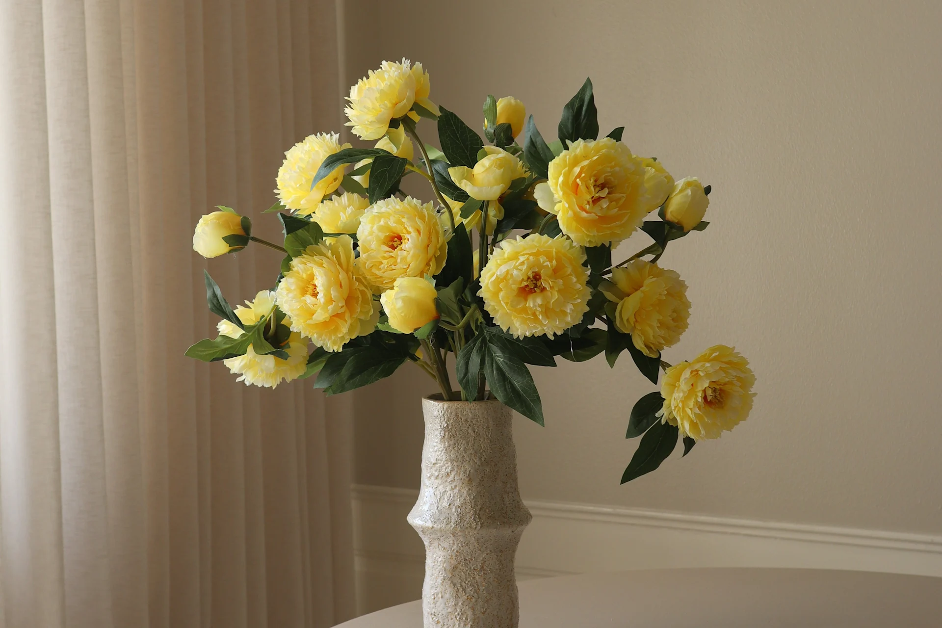 En lys, teksturert vase inneholder en livlig bukett med gule peonlignende blomster og grønne blader mot en lys bakgrunn.