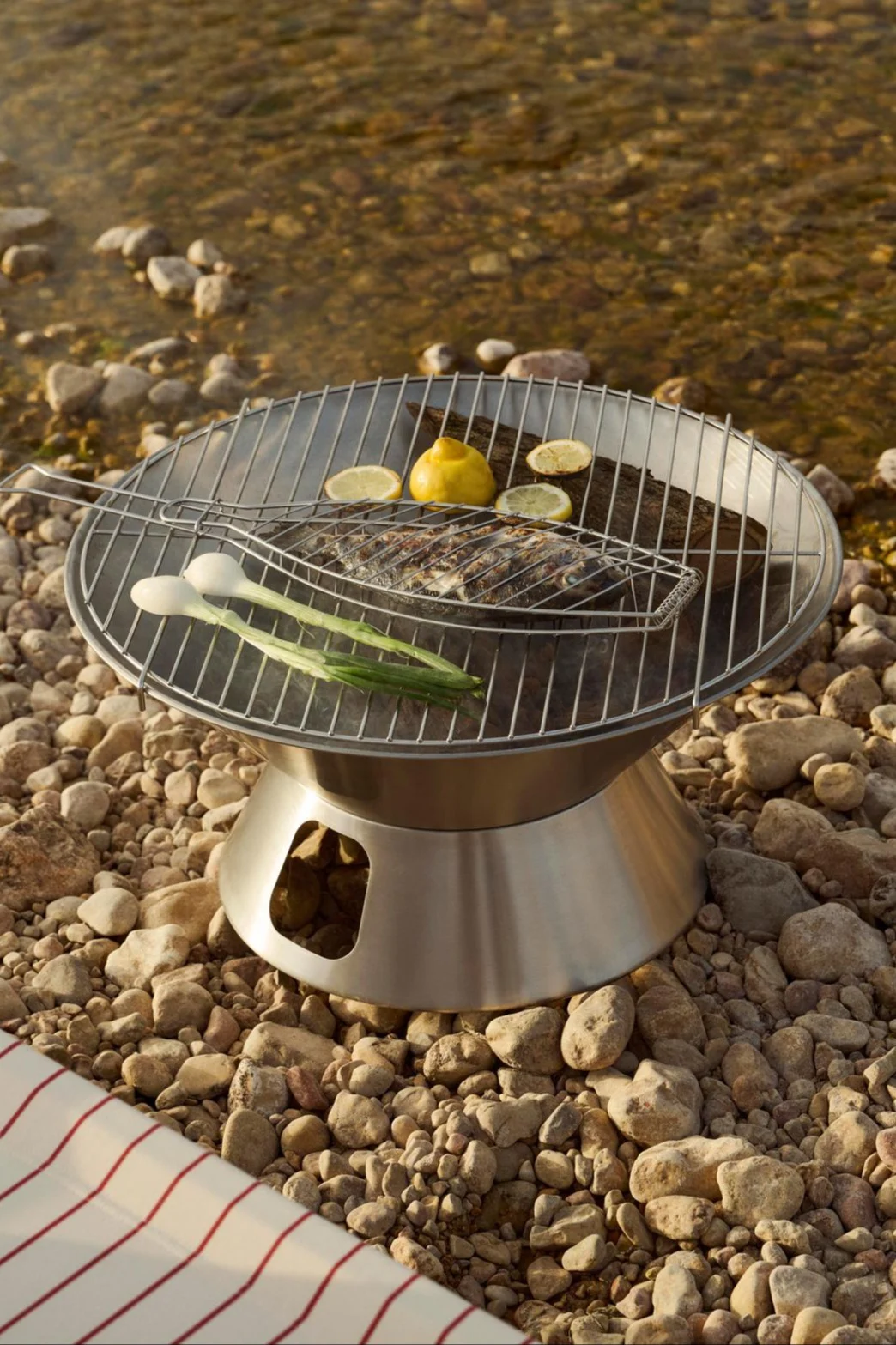En bærbar grill i rustfritt stål med en hel fisk i en grillkurv, sitroner og vårløk på en steinete elvebredd.