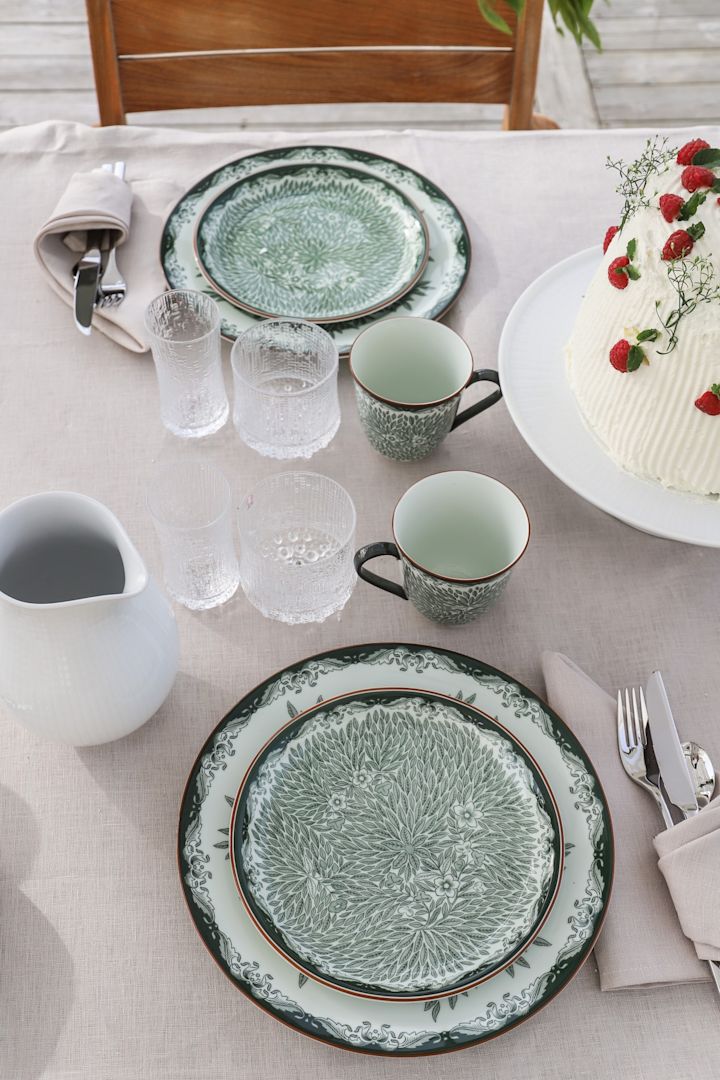 Tradisjonelle midtsommertips inkluderer selvfølgelig å spise ute i det fri. Her ser du det grønne serviset Ostindia og Ostindia Floris på bordet, sammen med Ultima Thule-glass fra Iittala.
