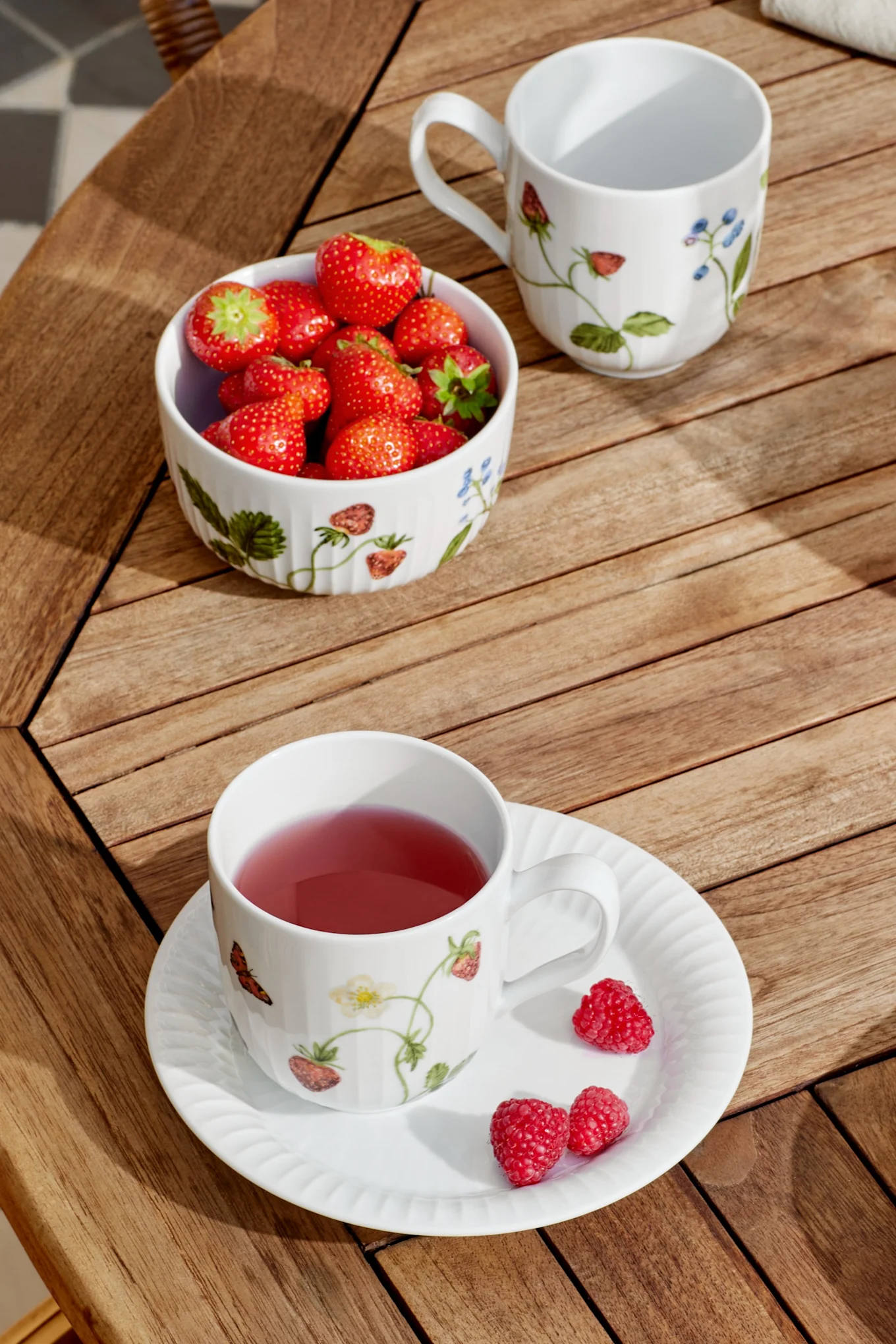 En hvit kopp med rosa te og et fat med bringebær, en bolle jordbær og en tom kopp står på et spilebord i tre i sollys.