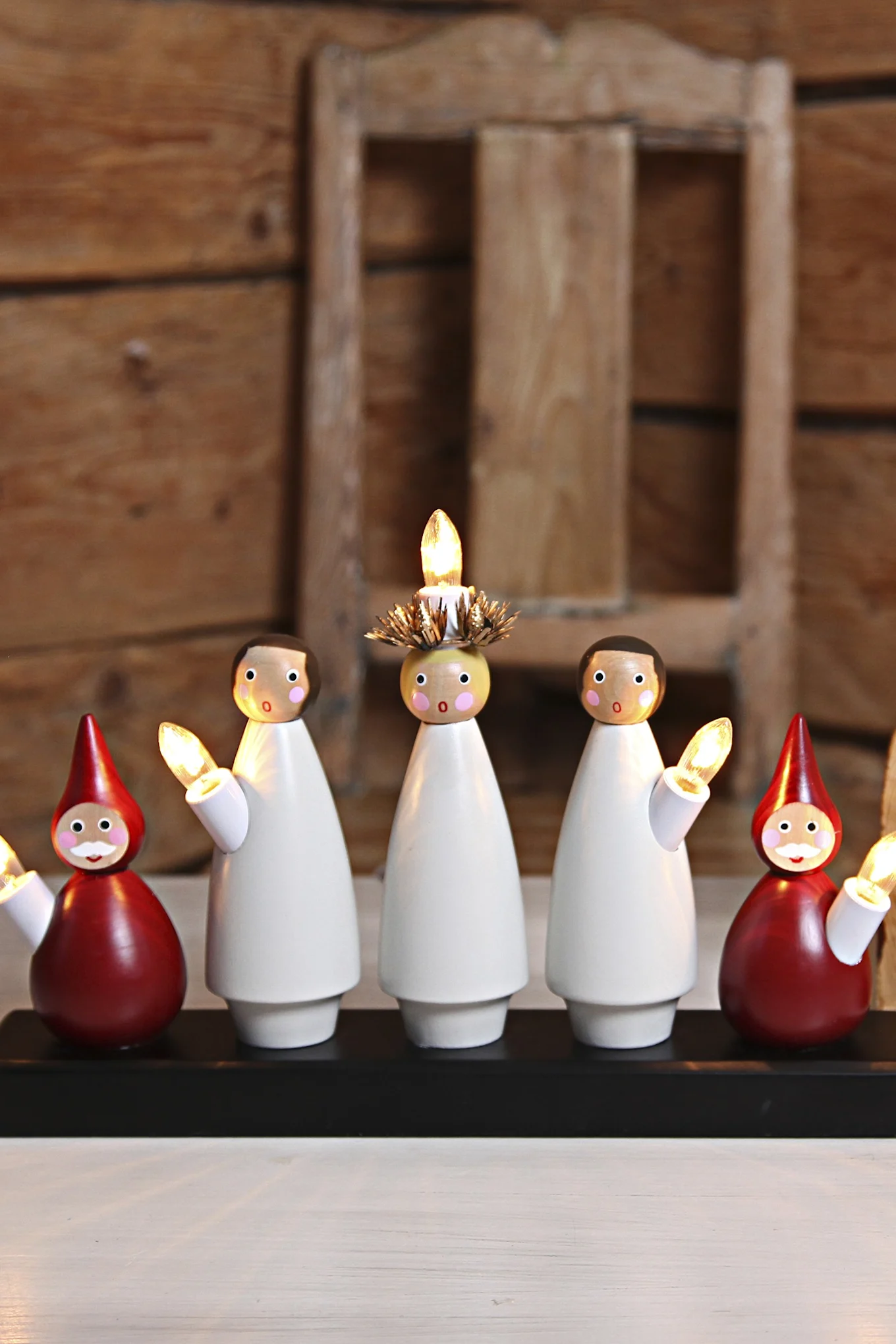 Fem hvite snømannfigurer i tre med glødende lys på en hvit base, ved siden av et juletre.