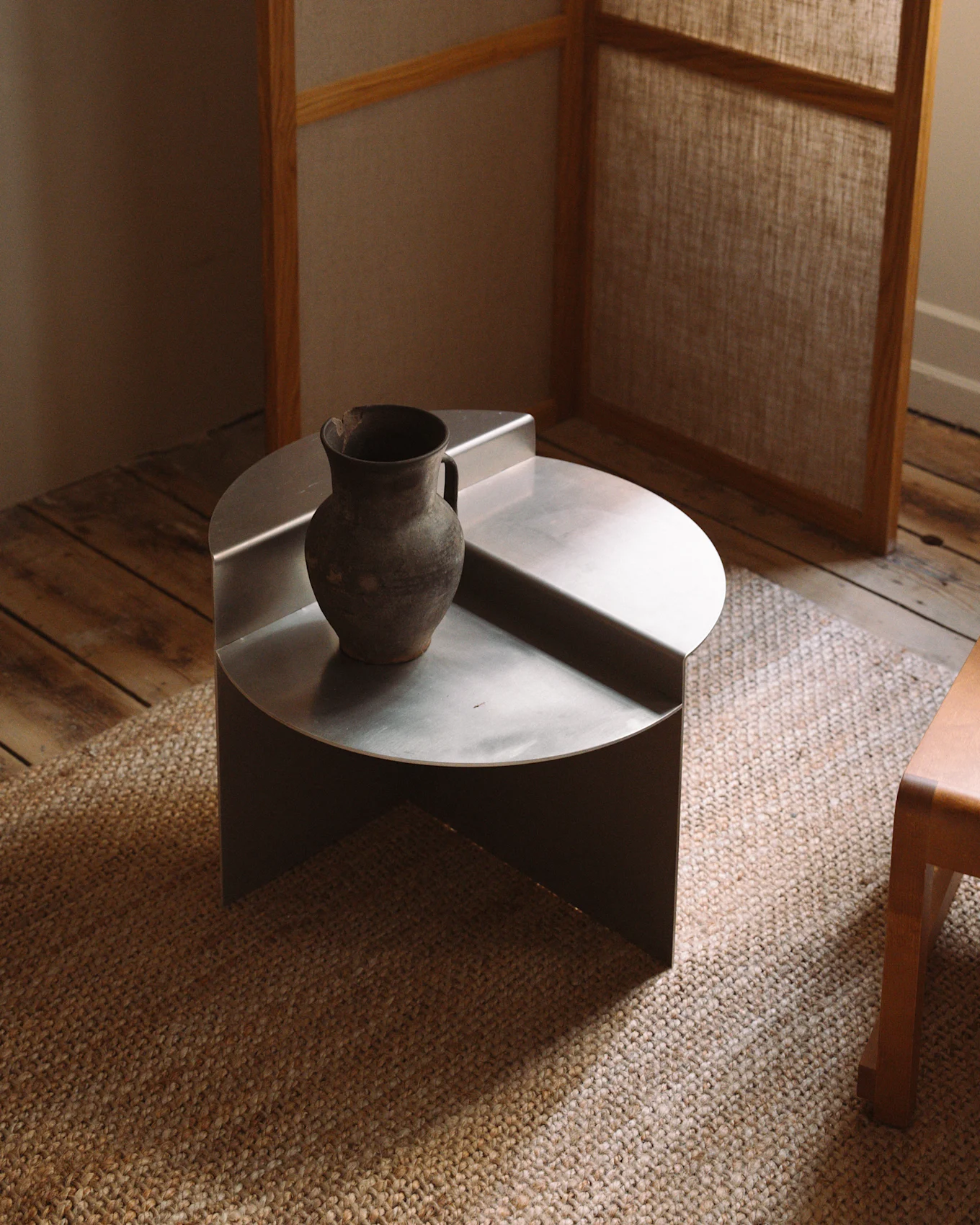 Et minimalistisk, lavt, rundt sofabord i sølvmetall med en rustikk mørk keramisk vase på et vevd teppe.