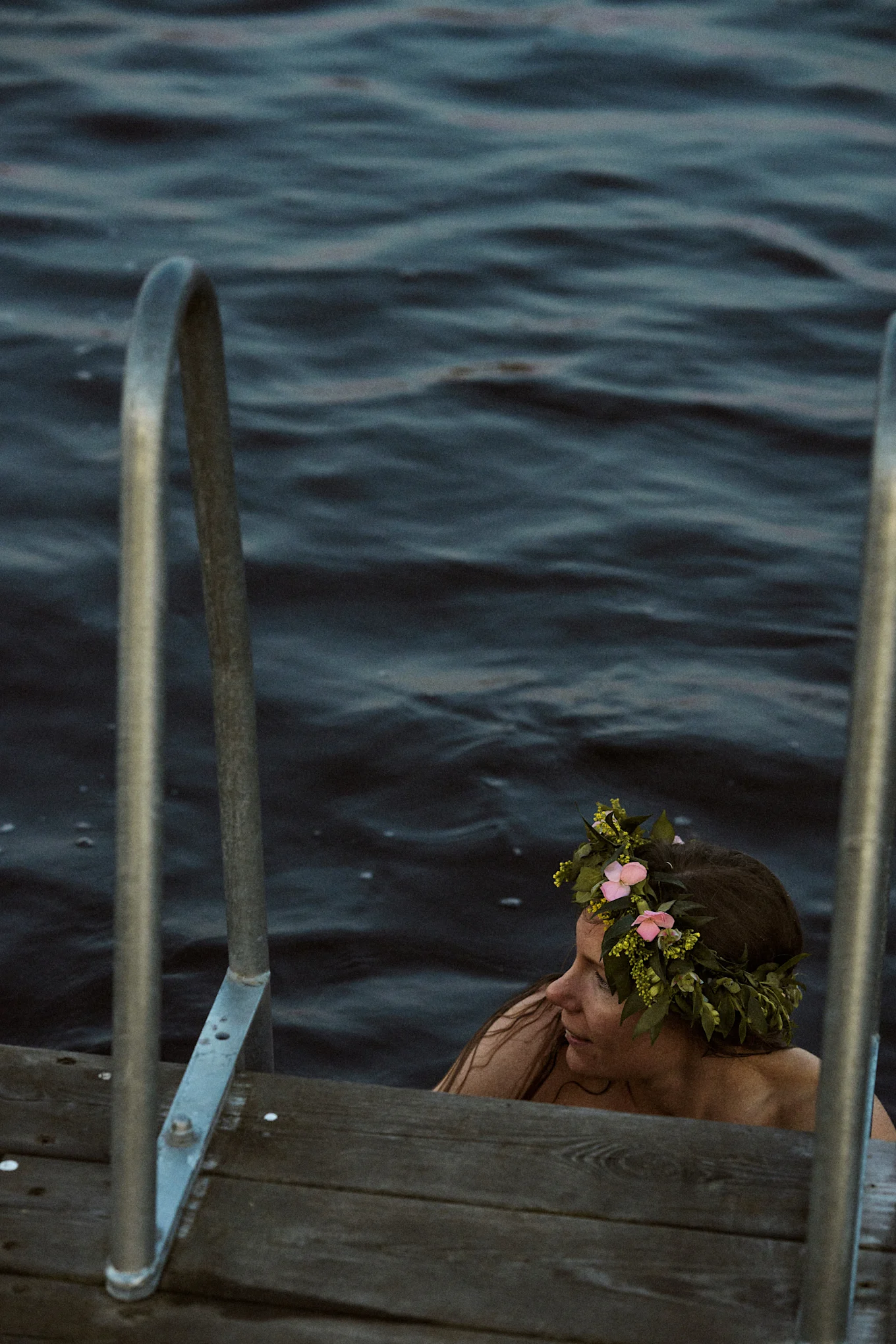 Kvinne i mørkt vann med blomsterkrans, ved siden av en trebrygge med metallrekkverk.
