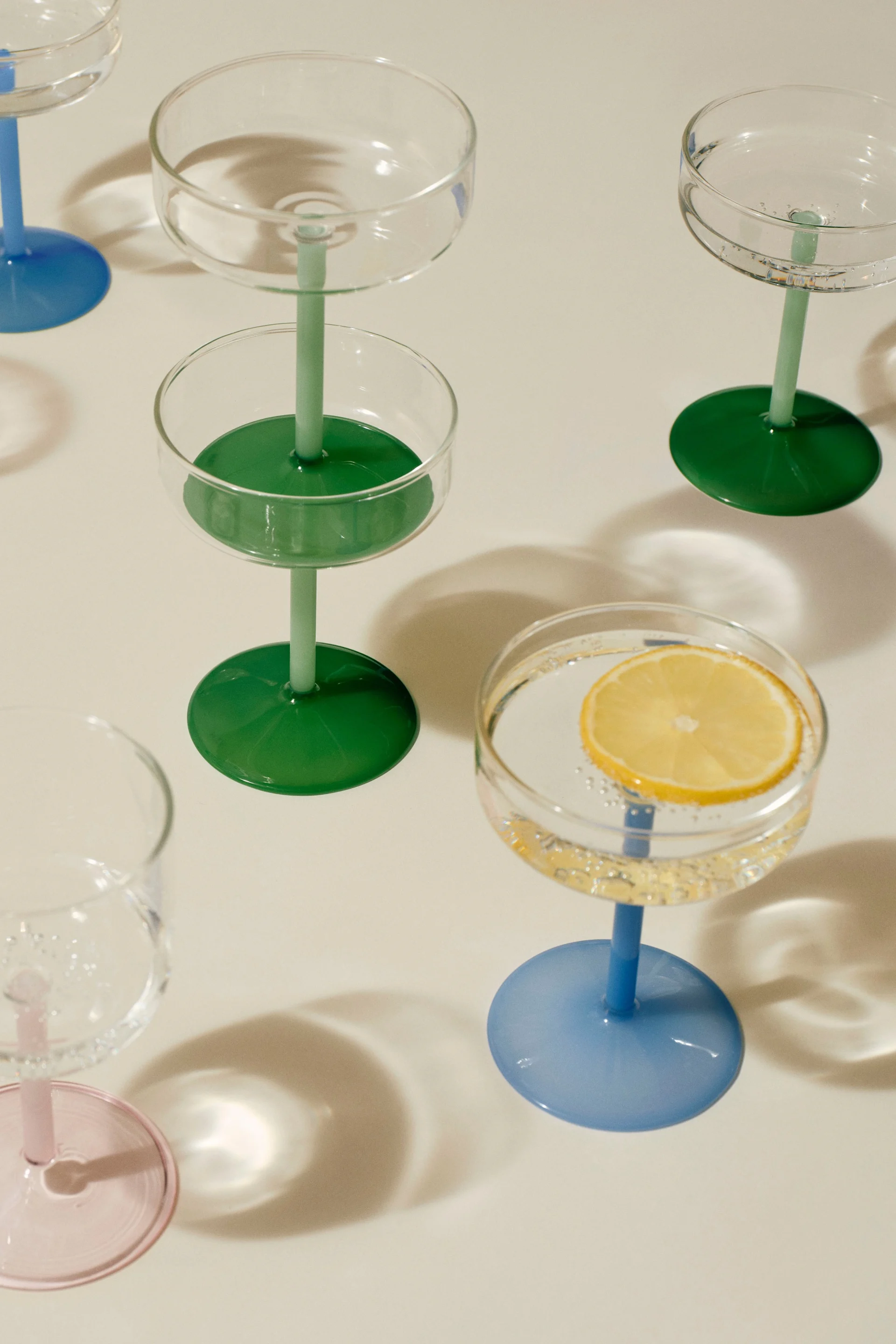 Utvalg av coupeglass med fargerike stett og baser. Ett glass inneholder en sitronskive i sprudlende vann.
